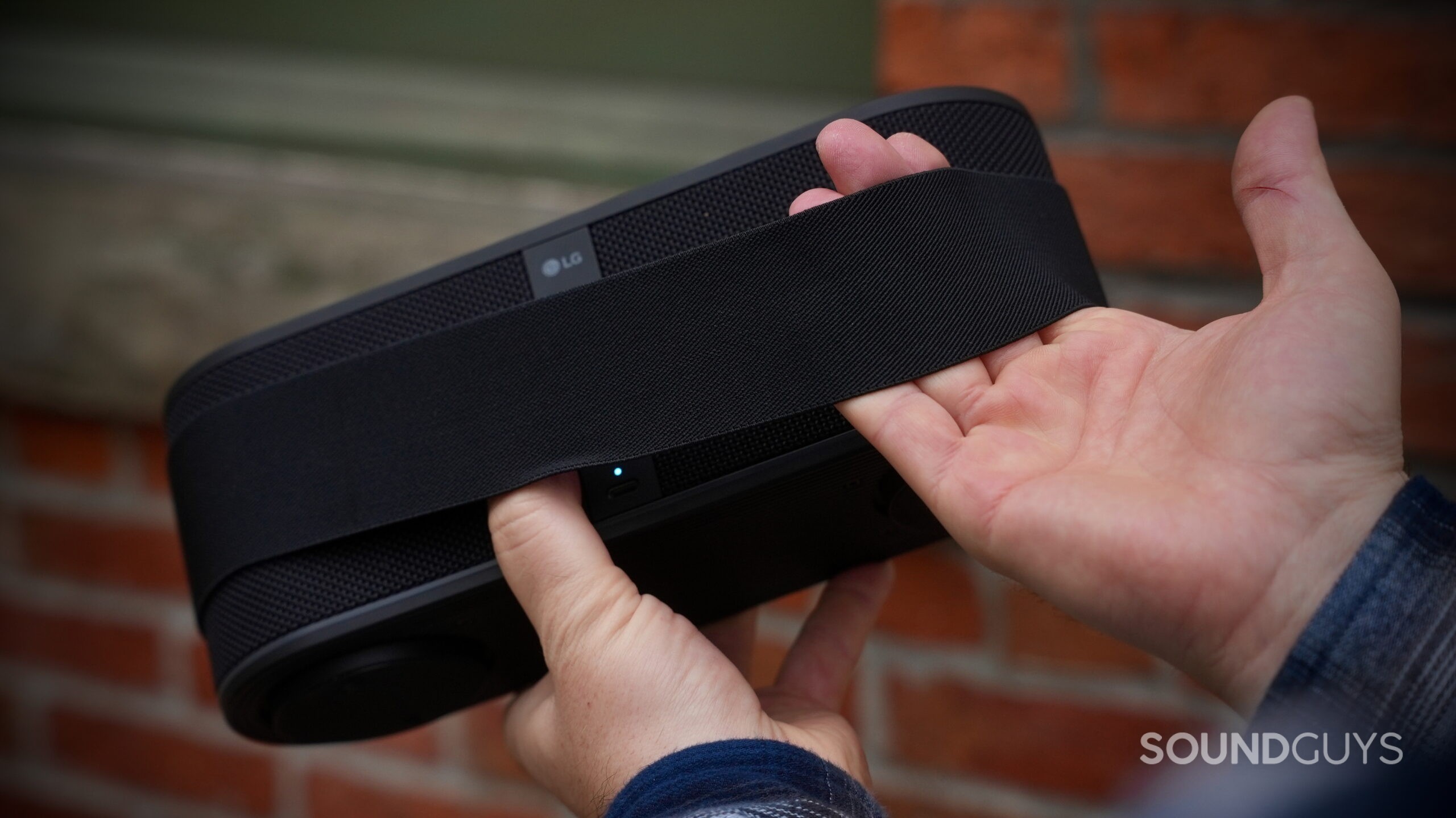 A hand holding the LG xboom Bounce speaker by the back, with its attachment strap wrapped around his hand.