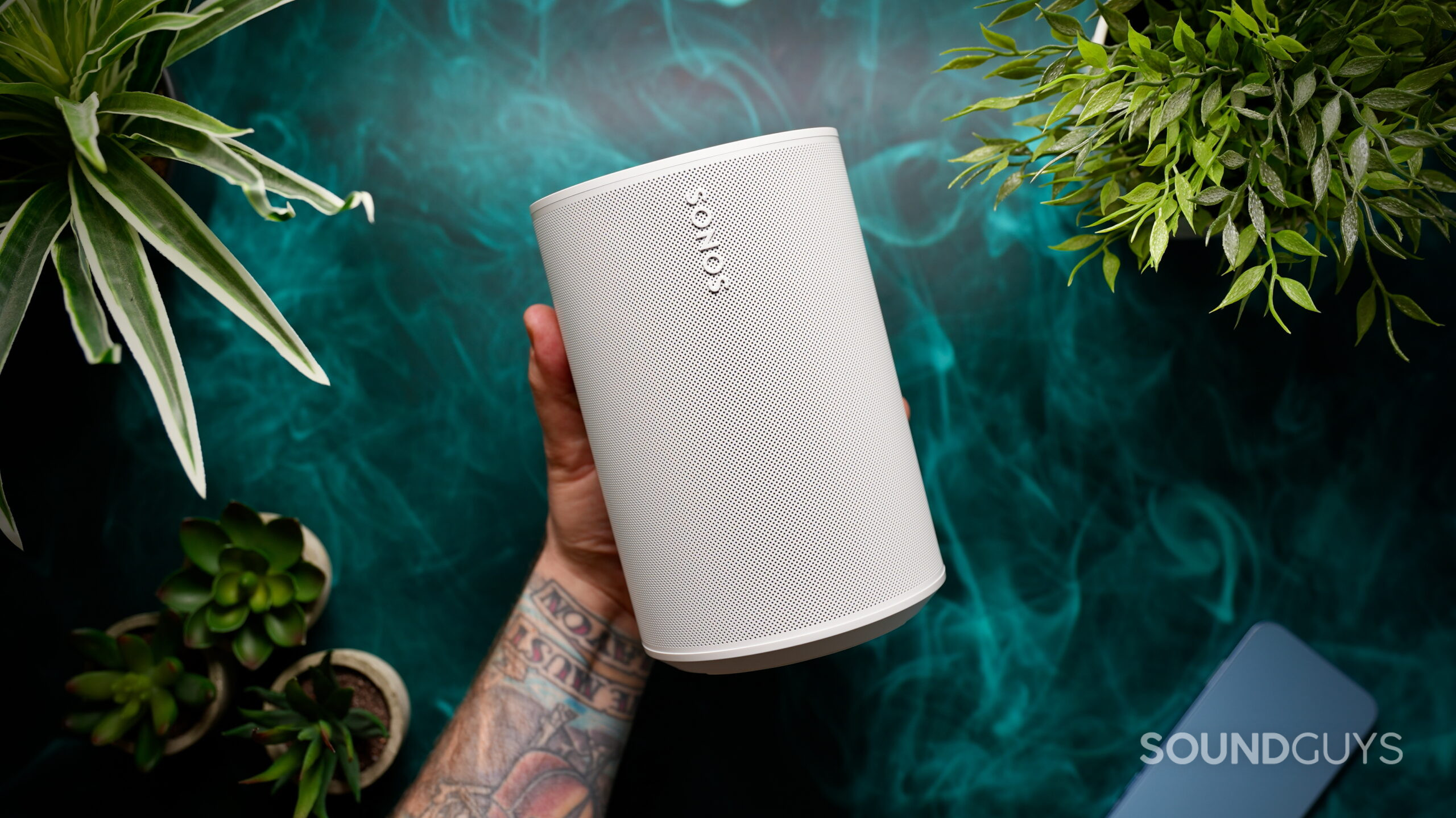 Sonos Era 100 SL - 1 Hand holding the Sonos Era 100 SL speaker in white against a green background with plants and a smartphone nearby.