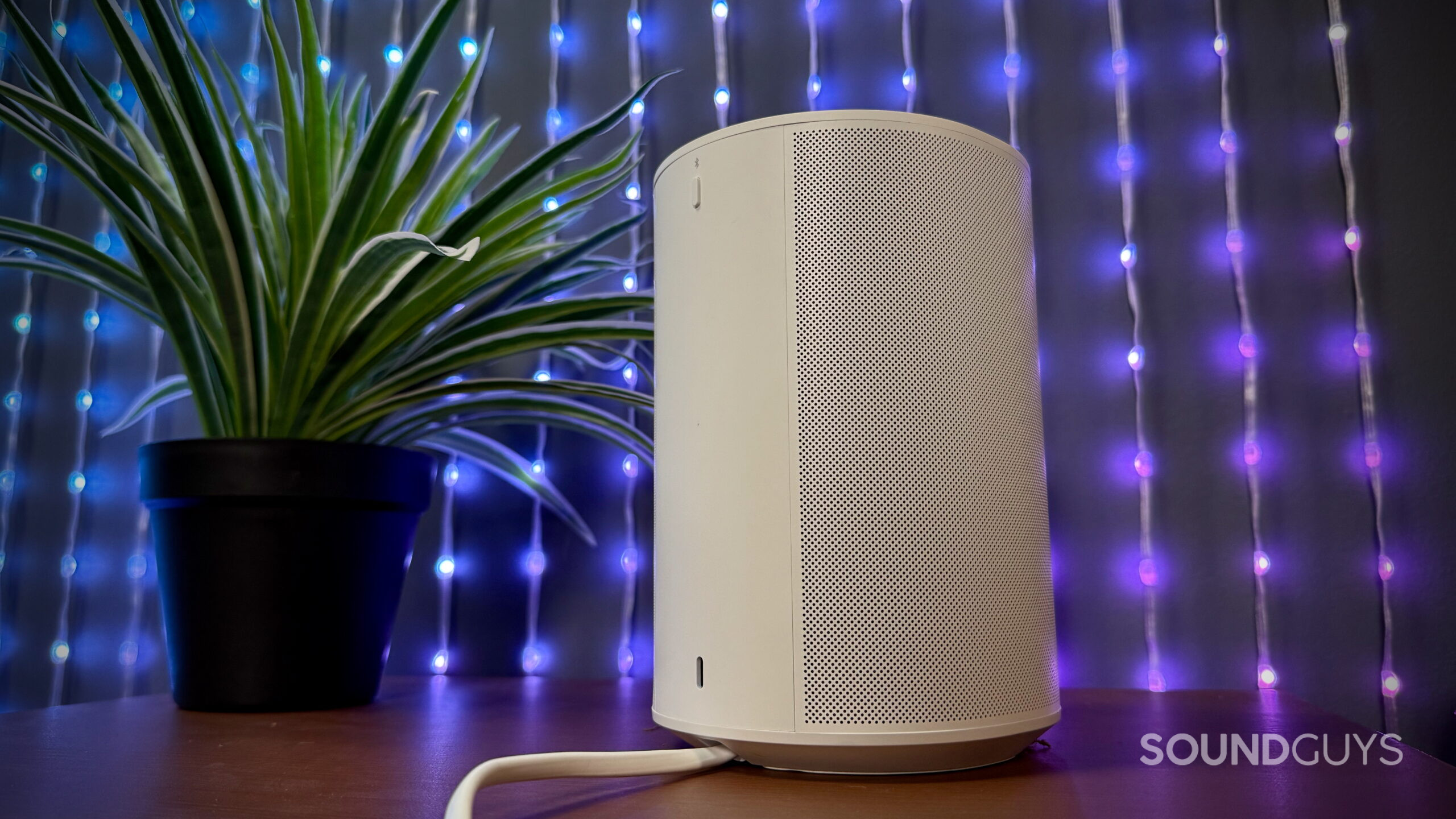 Sonos Era 100 SL - 6 Rear angle of the Sonos Era 100 SL speaker in white showing the perforated grille and power cable on a desk with decorative lights behind it.