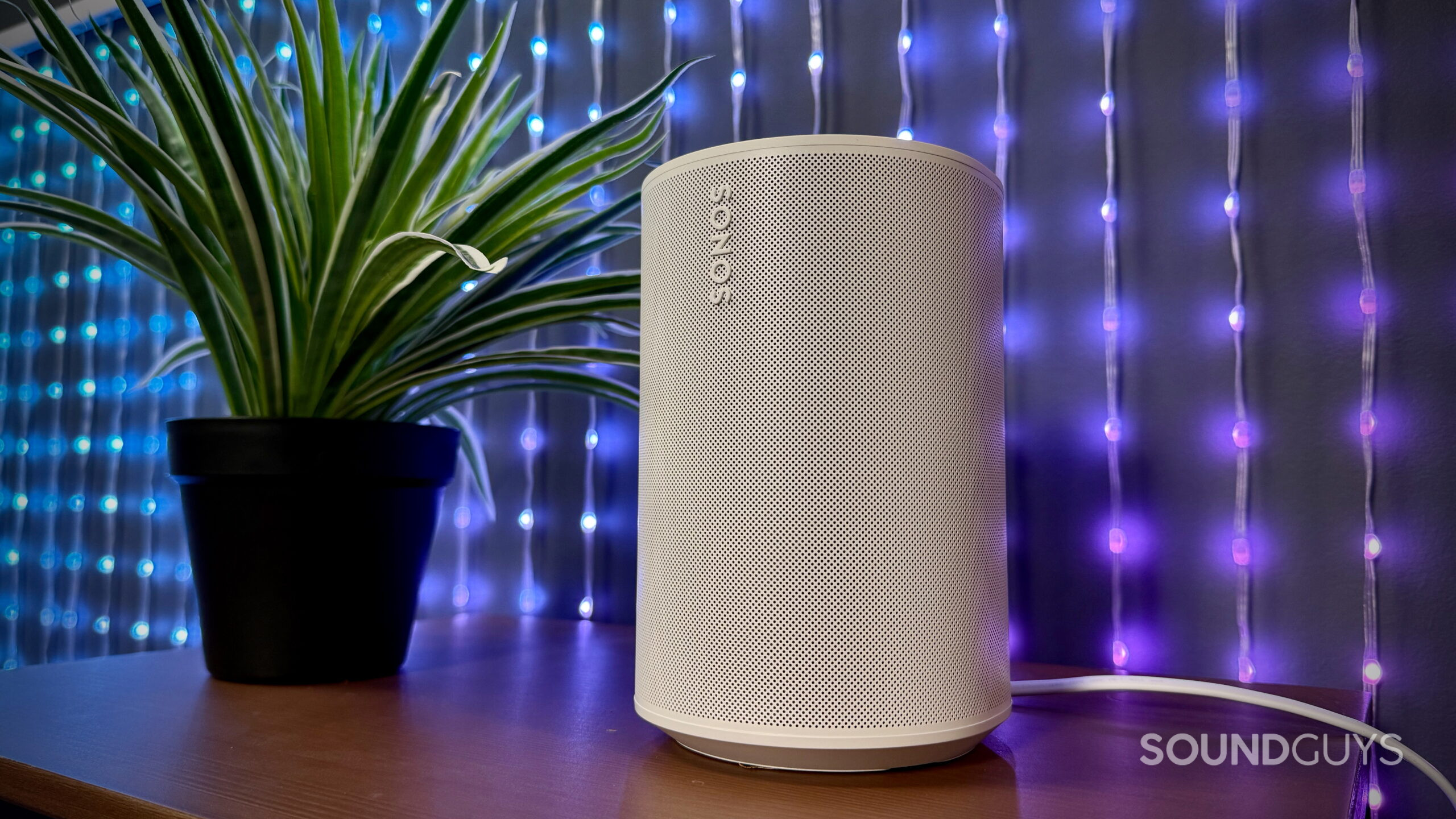 Sonos Era 100 SL - 7 Sonos Era 100 SL smart speaker in white on a wooden desk next to a potted plant with blue and purple LED lights in the background.