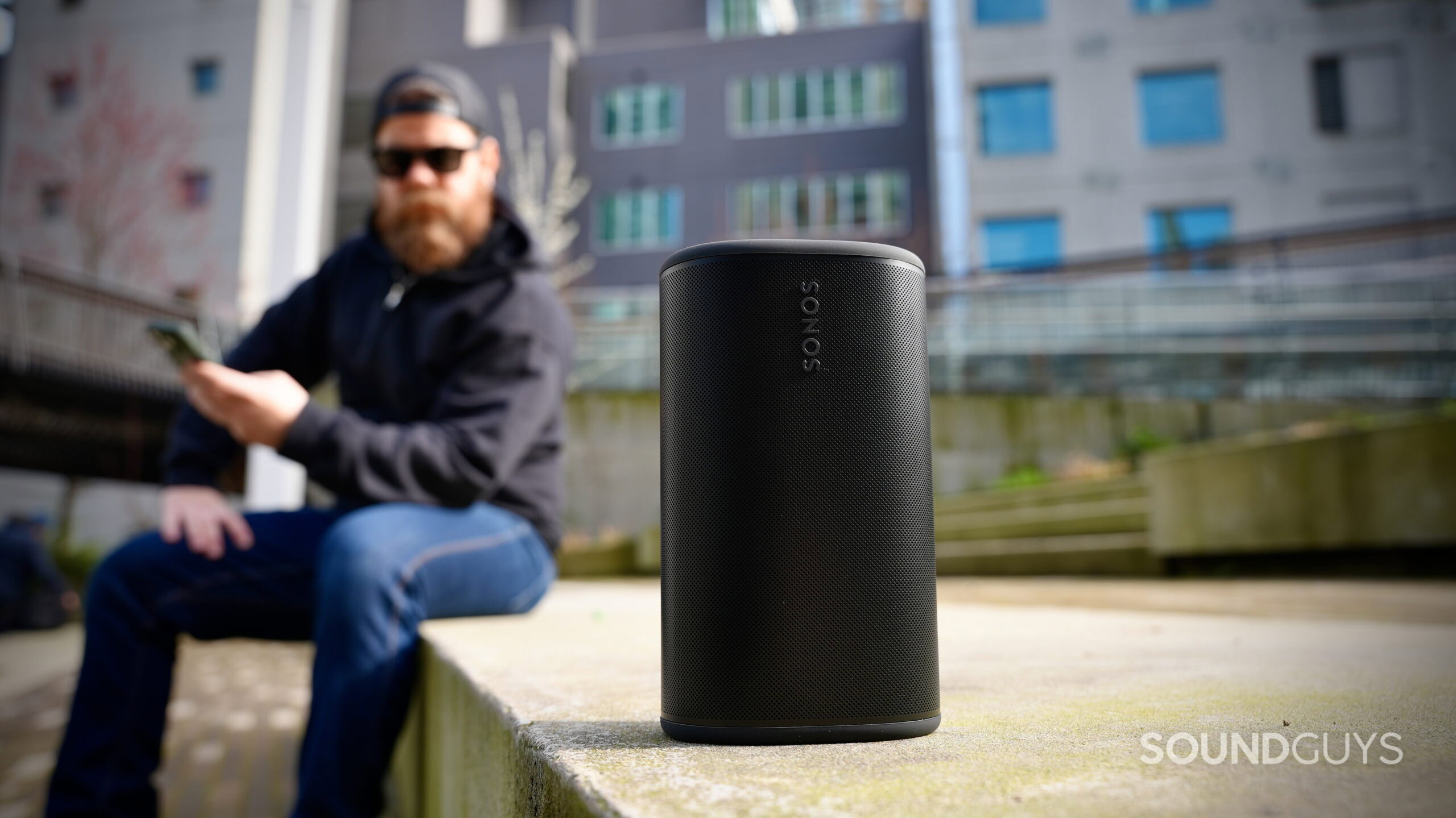 The Sonos Play portable speaker on a concrete ledge outdoors with a person sitting in the background.