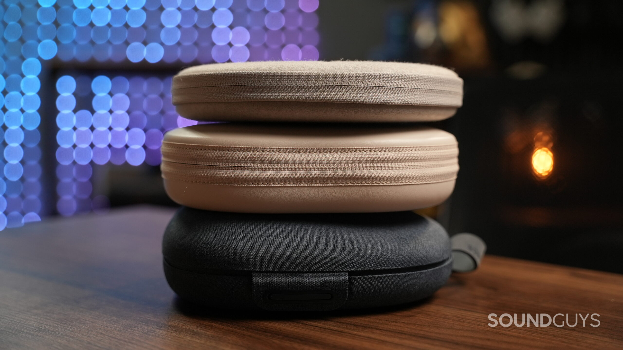 headphone cases stacked Three headphone carrying cases stacked on a wooden surface, showing different finishes and textures, with soft bokeh lights in the background.