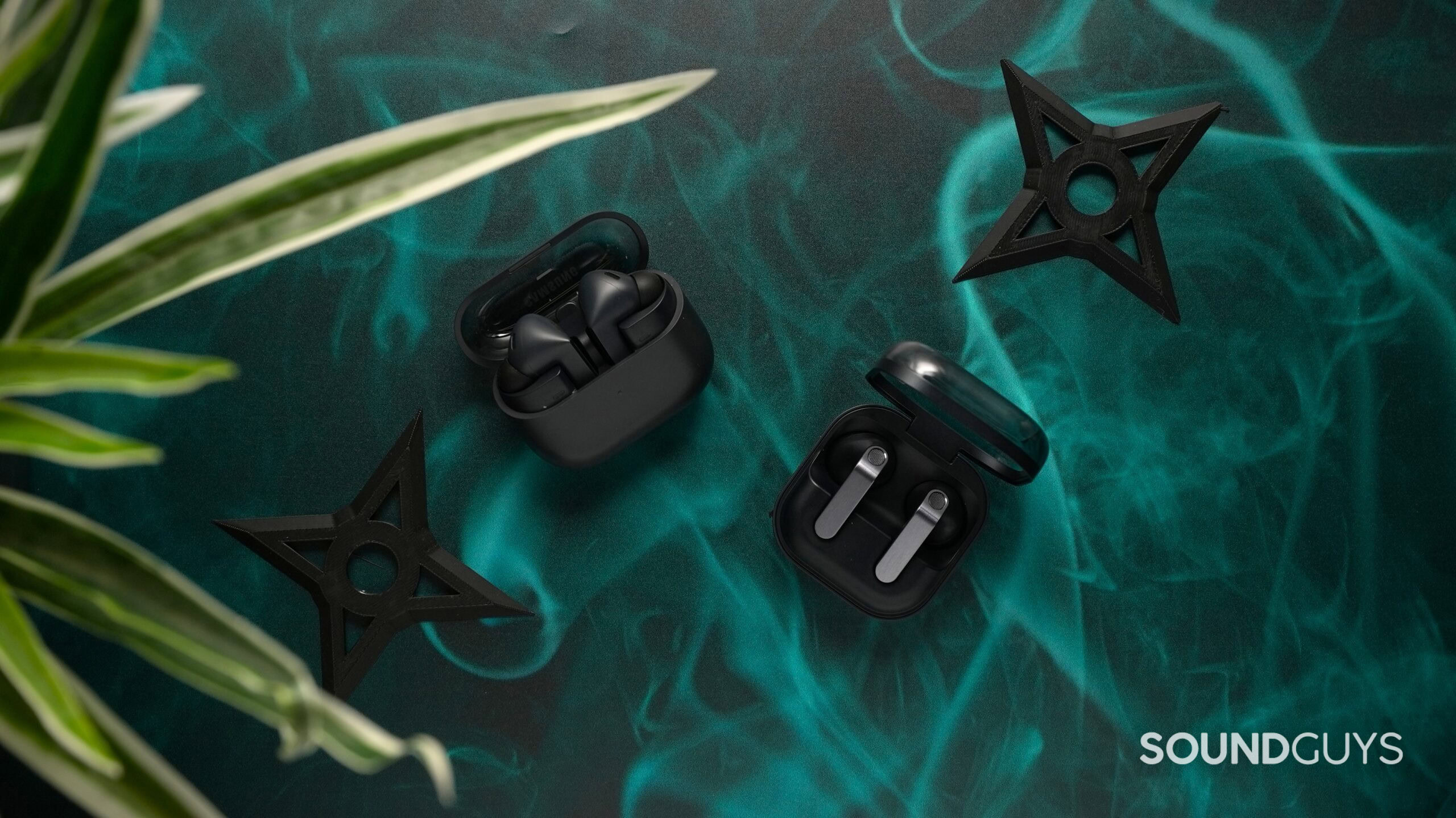 Top-down product shot of two wireless earbuds sets—the Samsung Galaxy Buds 3 FE and Galaxy Buds 4—displayed in open charging cases on a teal, smoky background with decorative black shuriken shapes and green plant leaves framing the scene.