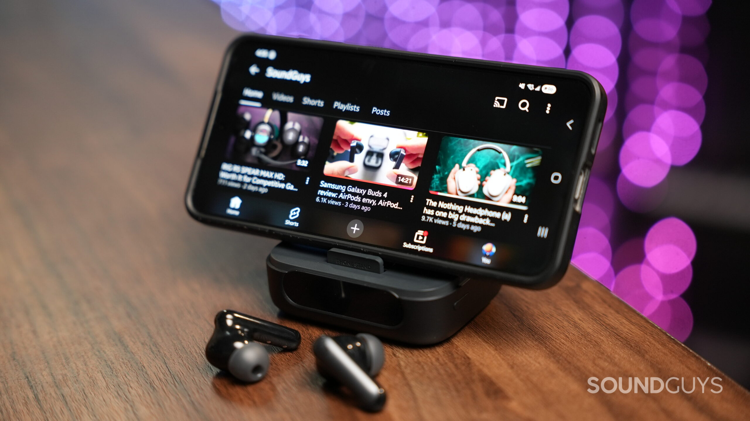 Soundcore P40i earbuds on a wooden table next to a smartphone displaying the SoundGuys YouTube channel.