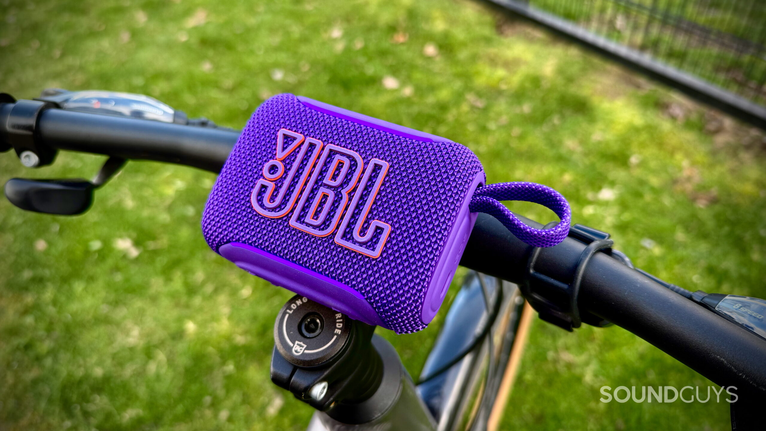 JBL Go 5 resting on bicycle handlebars over a grassy park background