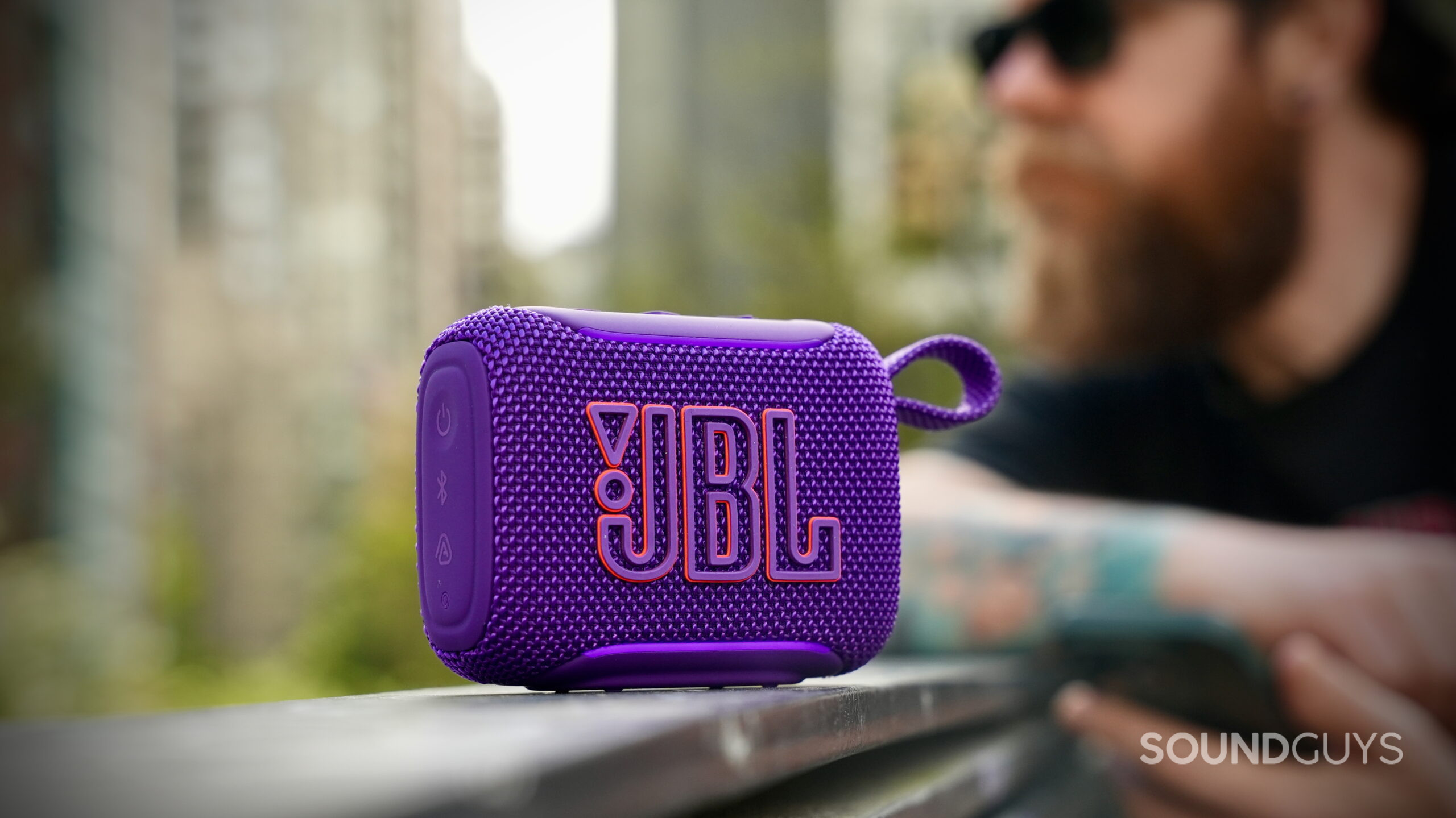 JBL Go 5 purple speaker on a railing with a person blurred in the background.