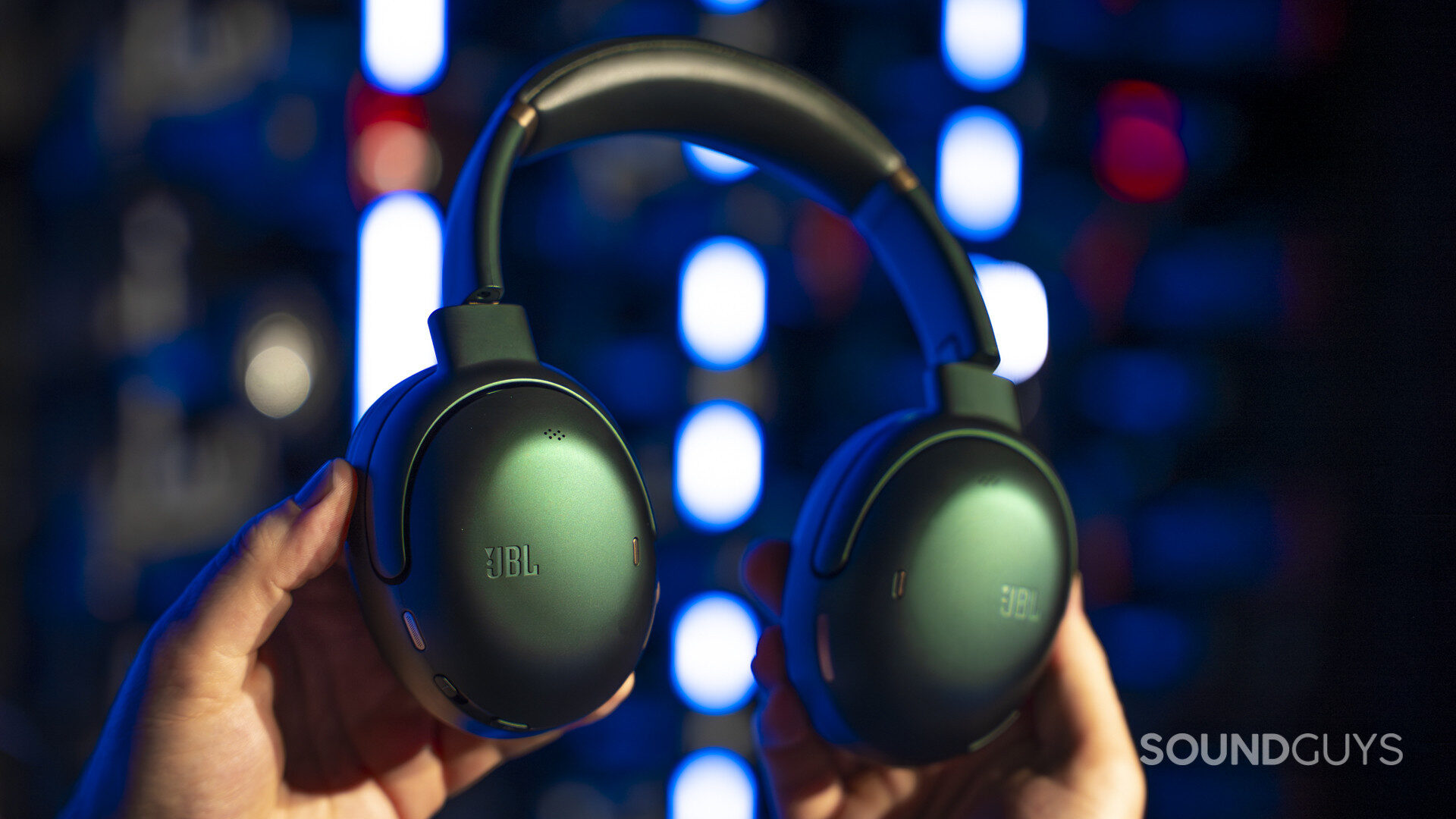 JBL Tour One M3 - 80th Anniversary Edition-1 Hands holding green JBL over-ear headphones against a blurred background with colorful lights, highlighting both ear cups and headband.