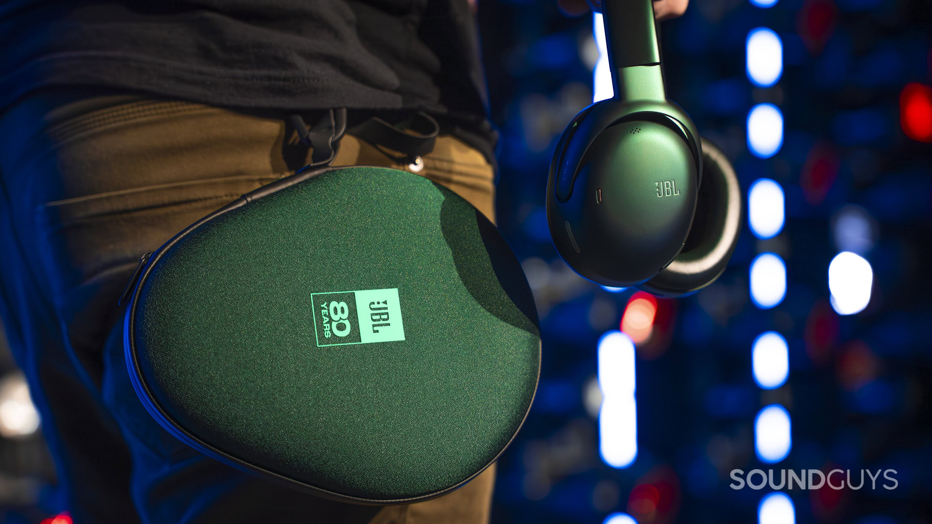 JBL Tour One M3 - 80th Anniversary Edition 2 Years Blue JBL headphones hang from the side of the same zippered carrying case with the “JBL 80 Years” logo, clipped to the person's waist.