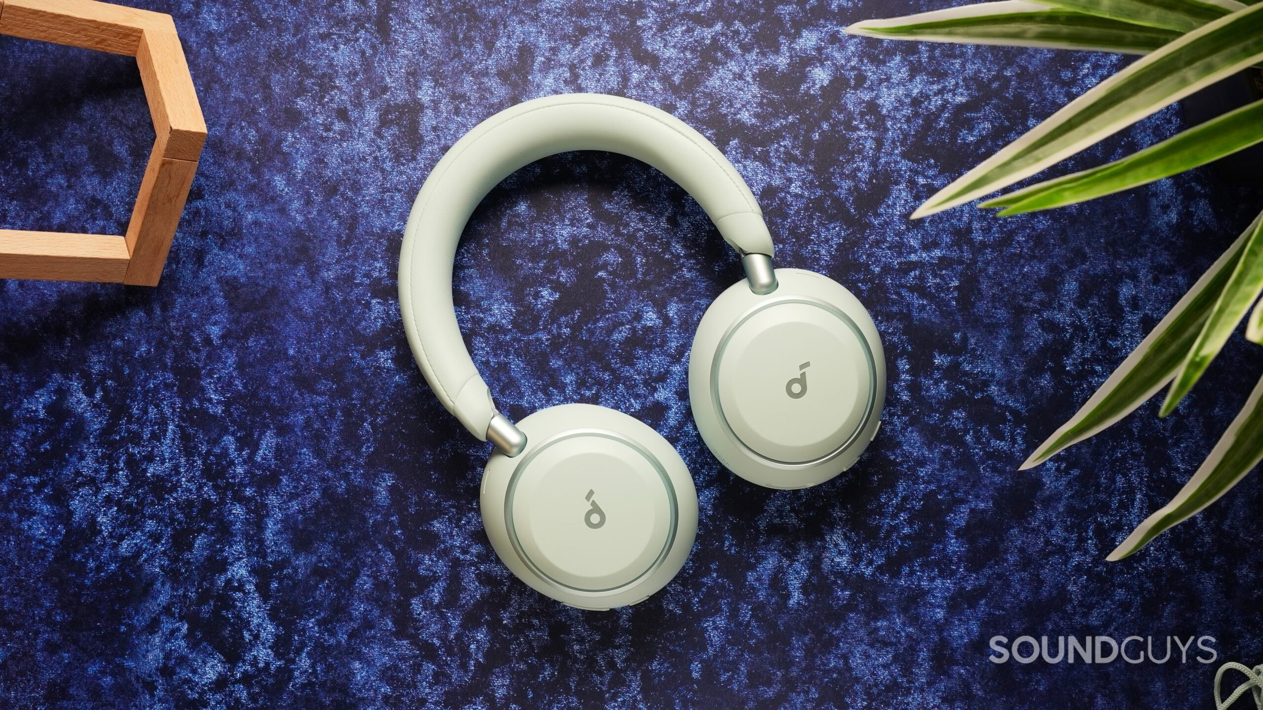 anker soundcore space 2 Light green Anker Soundcore Space 2 over-ear headphones laid flat on a textured blue surface, with a plant leaf and wooden decor in the frame.