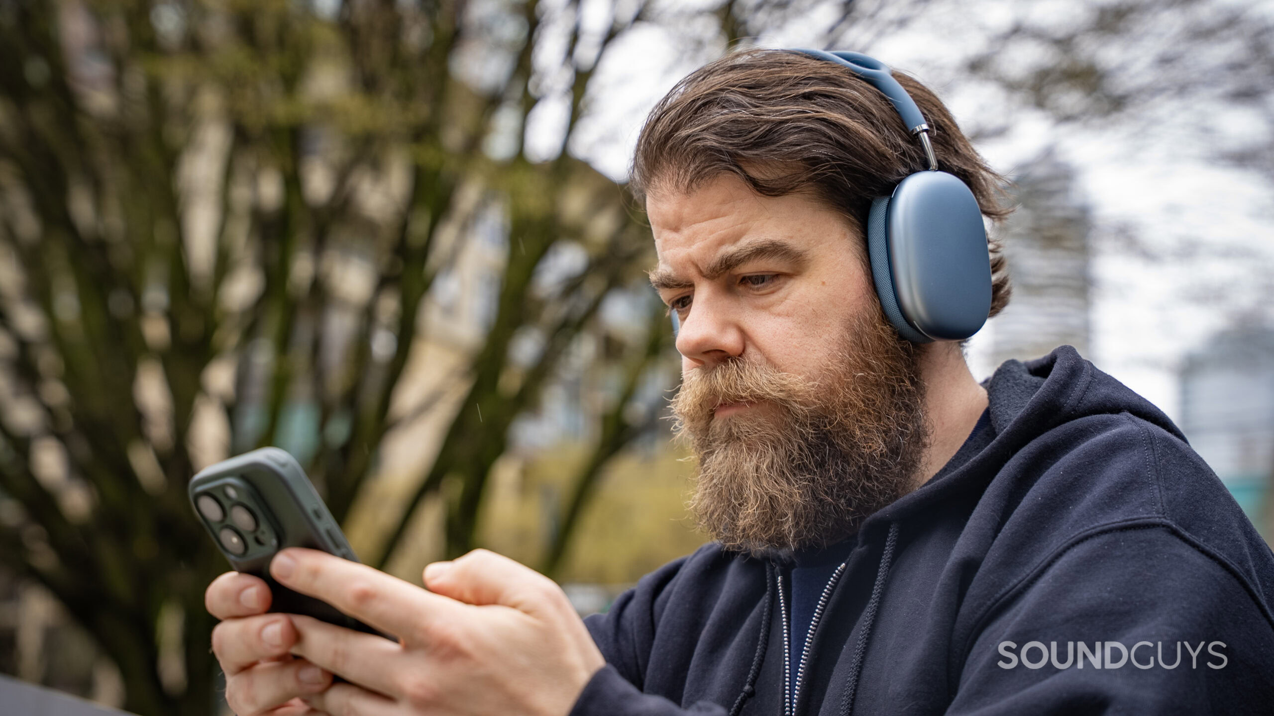 A man looks down using the iPhone 15 Pro in his hand while wearing the Apple AirPods Max 2 headphones