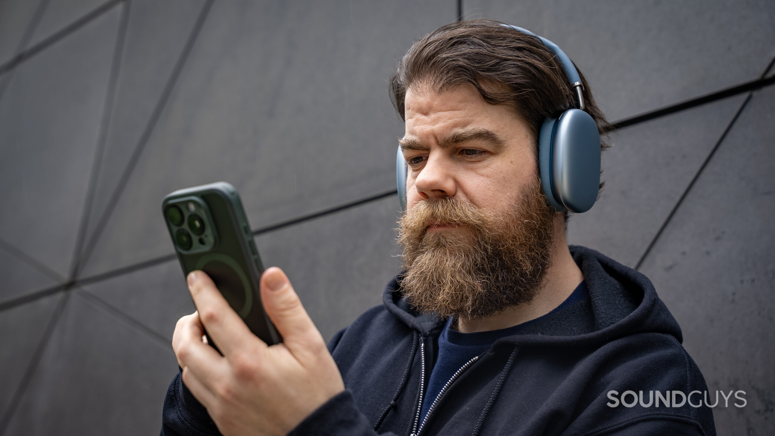 apple airpods max 2 phone in hand from side A man holding an iPhone 15 Pro in his hand while wearing the Apple AirPods Max 2 headphones