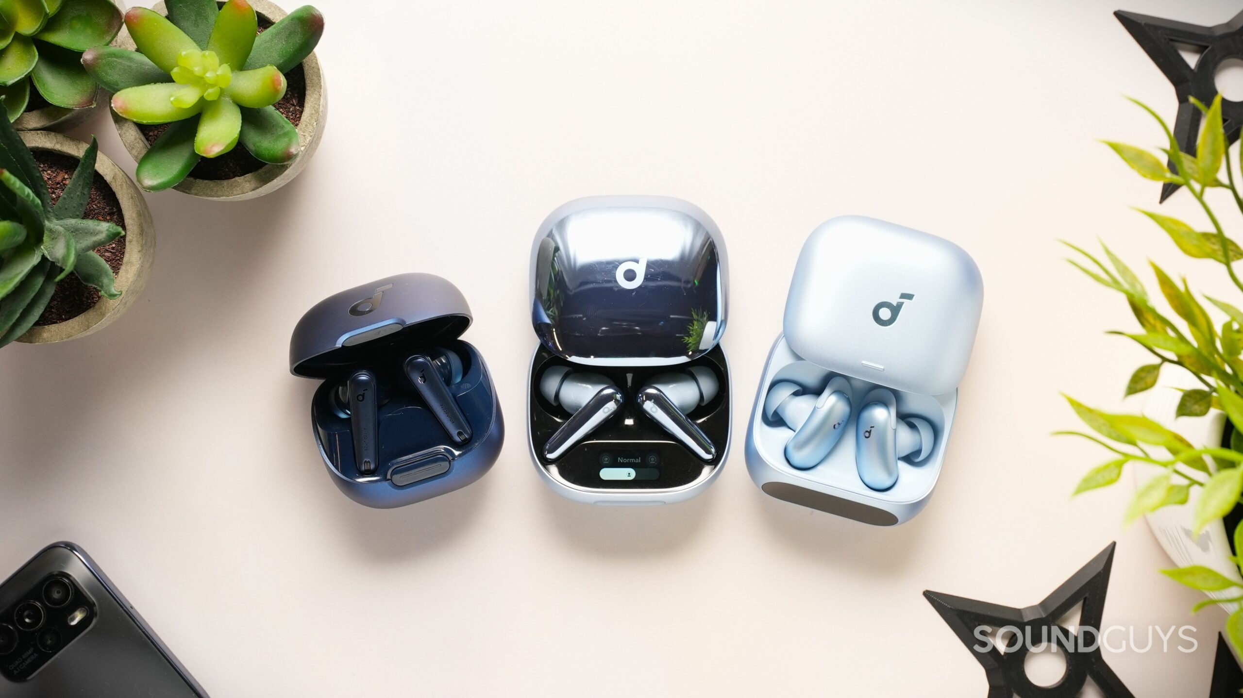 Three pairs of Soundcore wireless earbuds in open charging cases are arranged on a cream-colored desk with plants and black shuriken decorations around them. The earbuds feature dark blue, glossy silver, and matte light-blue finishes, photographed in a bright overhead setup.