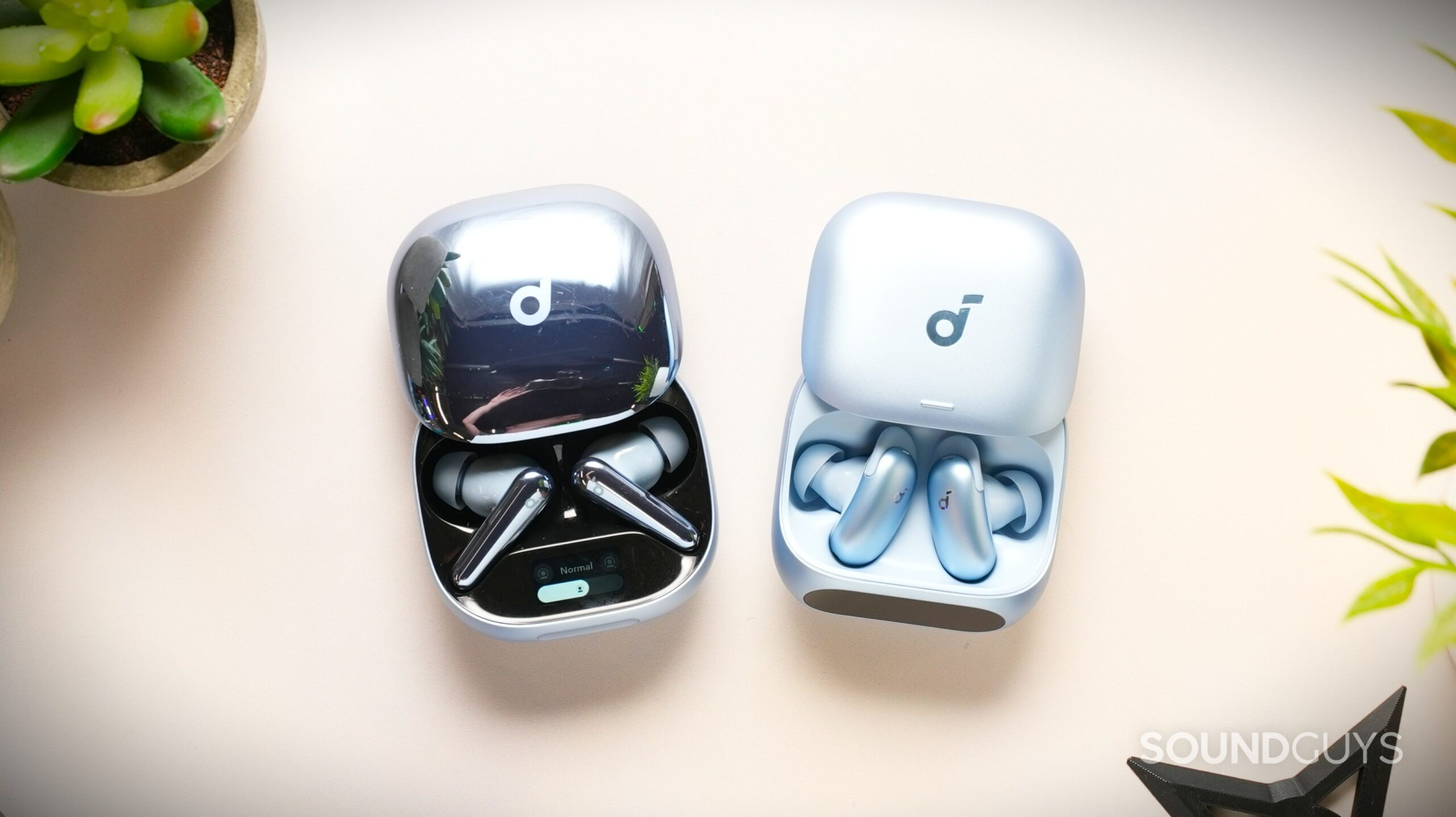 Two open light-blue and silver Soundcore wireless earbud charging cases sit side by side on a cream-colored desk, surrounded by small potted plants. The glossy dark silver earbuds on the left contrast with the matte light-blue earbuds on the right.