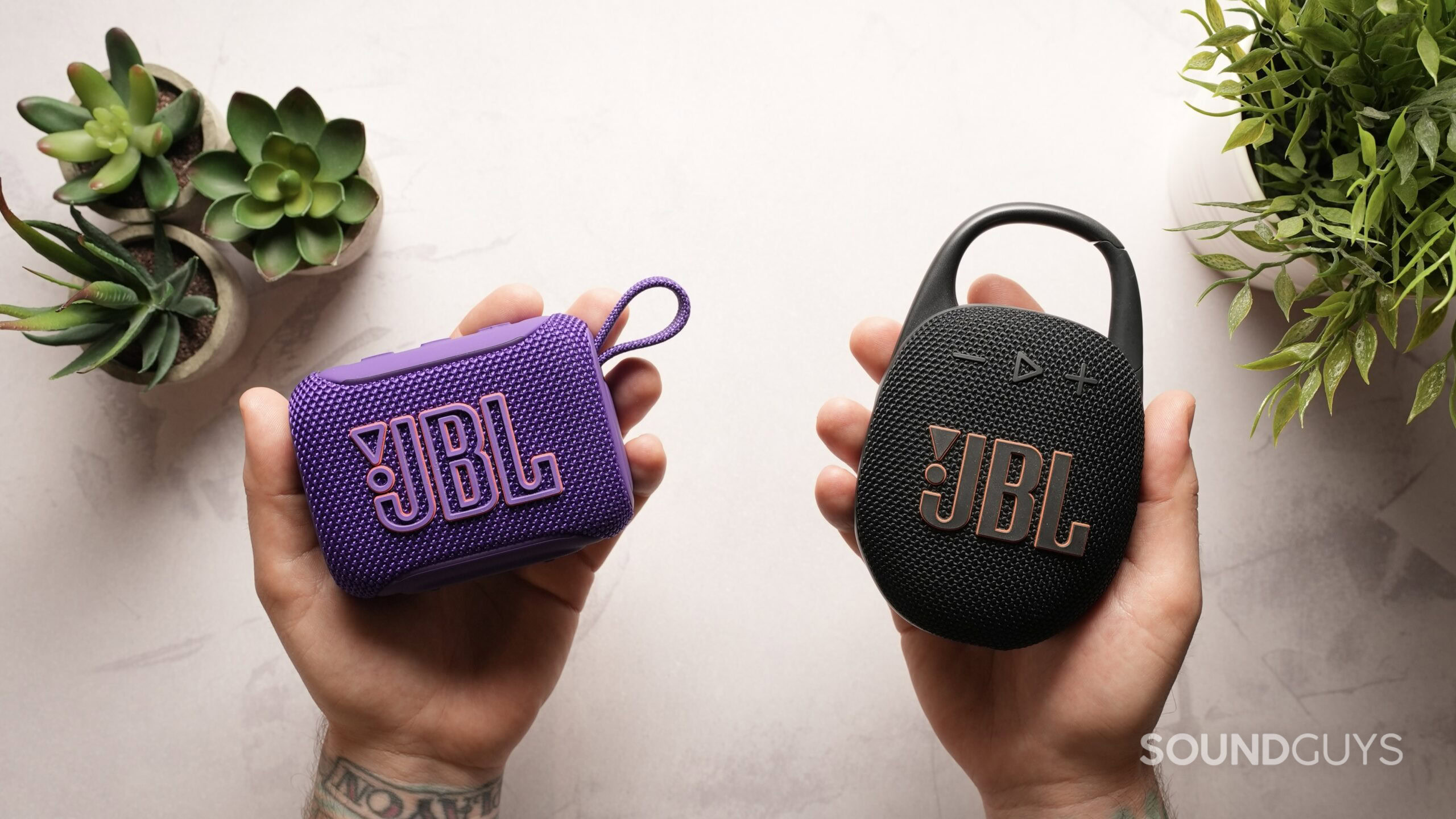 Hand holding the JBL Go 5 and JBL Clip 5 upright to compare their side profiles and onboard control button layouts.