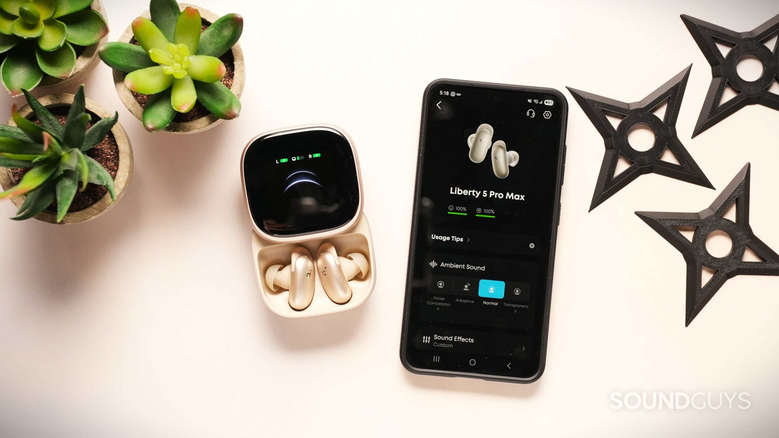 soundcore liberty 5 pro max with app on phone Soundcore Liberty 5 Pro Max earbuds beside a smartphone showing the companion app with ambient sound controls and battery status.