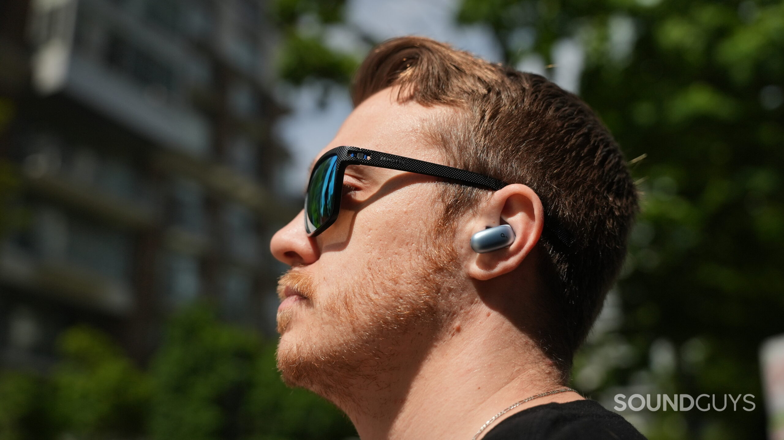 Outdoor portrait of a person wearing blue Soundcore Liberty 5 Pro Max earbuds and reflective sunglasses in bright sunlight.