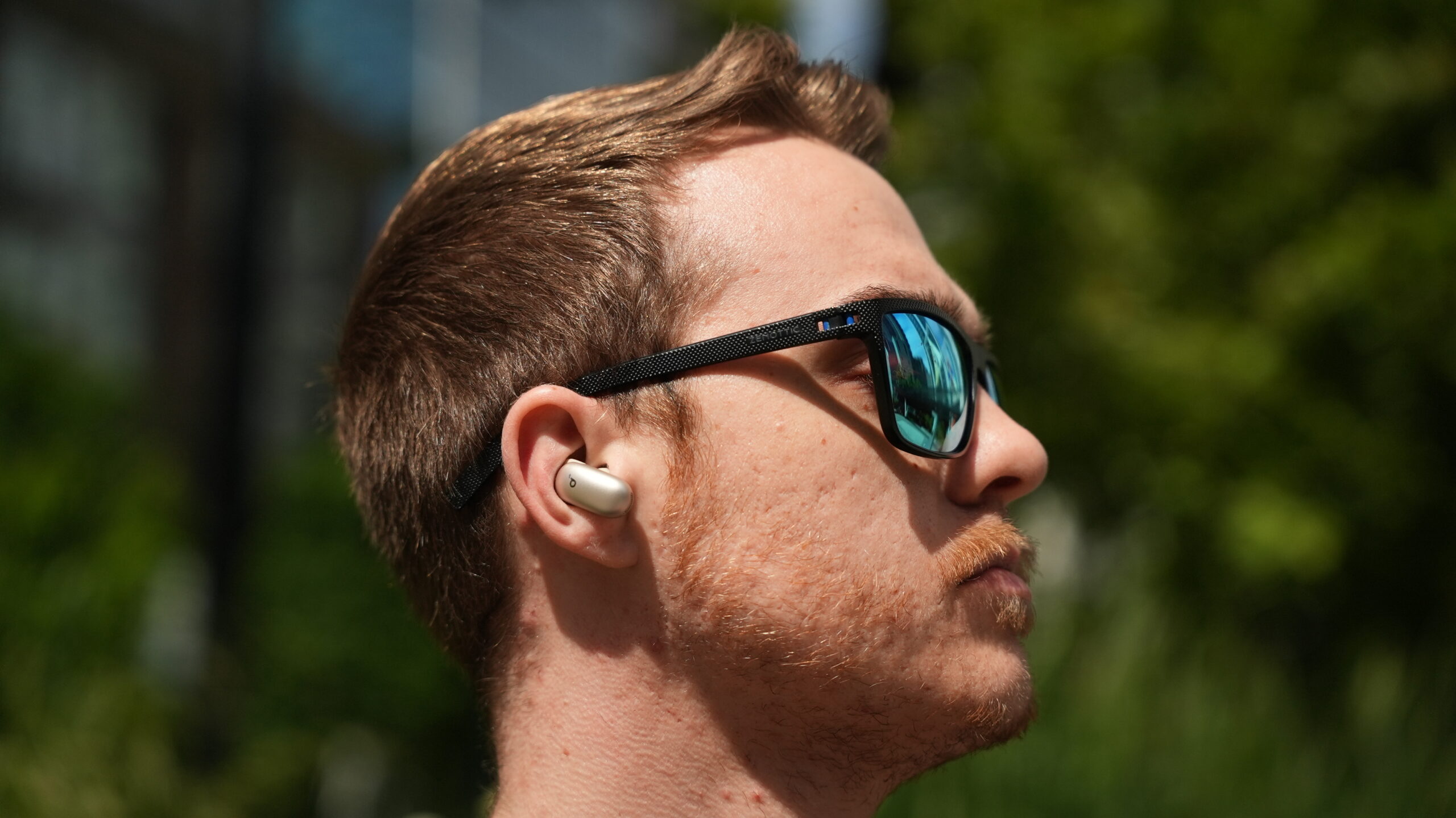 wearing the soundcore liberty 5 pro max Side profile of a person wearing beige Soundcore Liberty 5 Pro Max earbuds and black sunglasses outdoors in daylight.