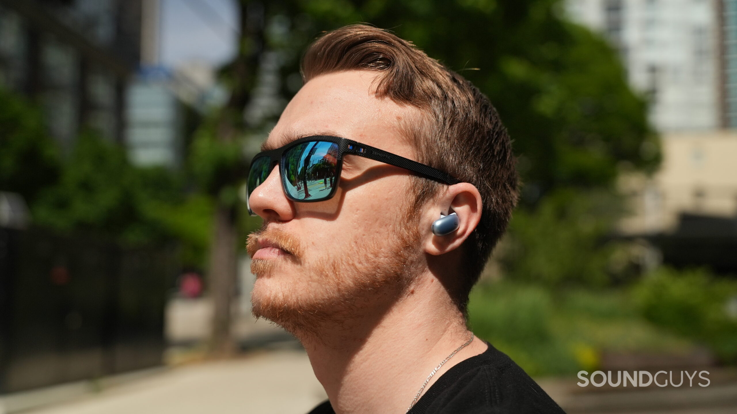 Outdoor lifestyle shot of a person wearing blue Soundcore Liberty 5 Pro earbuds with reflective sunglasses against a blurred city background.