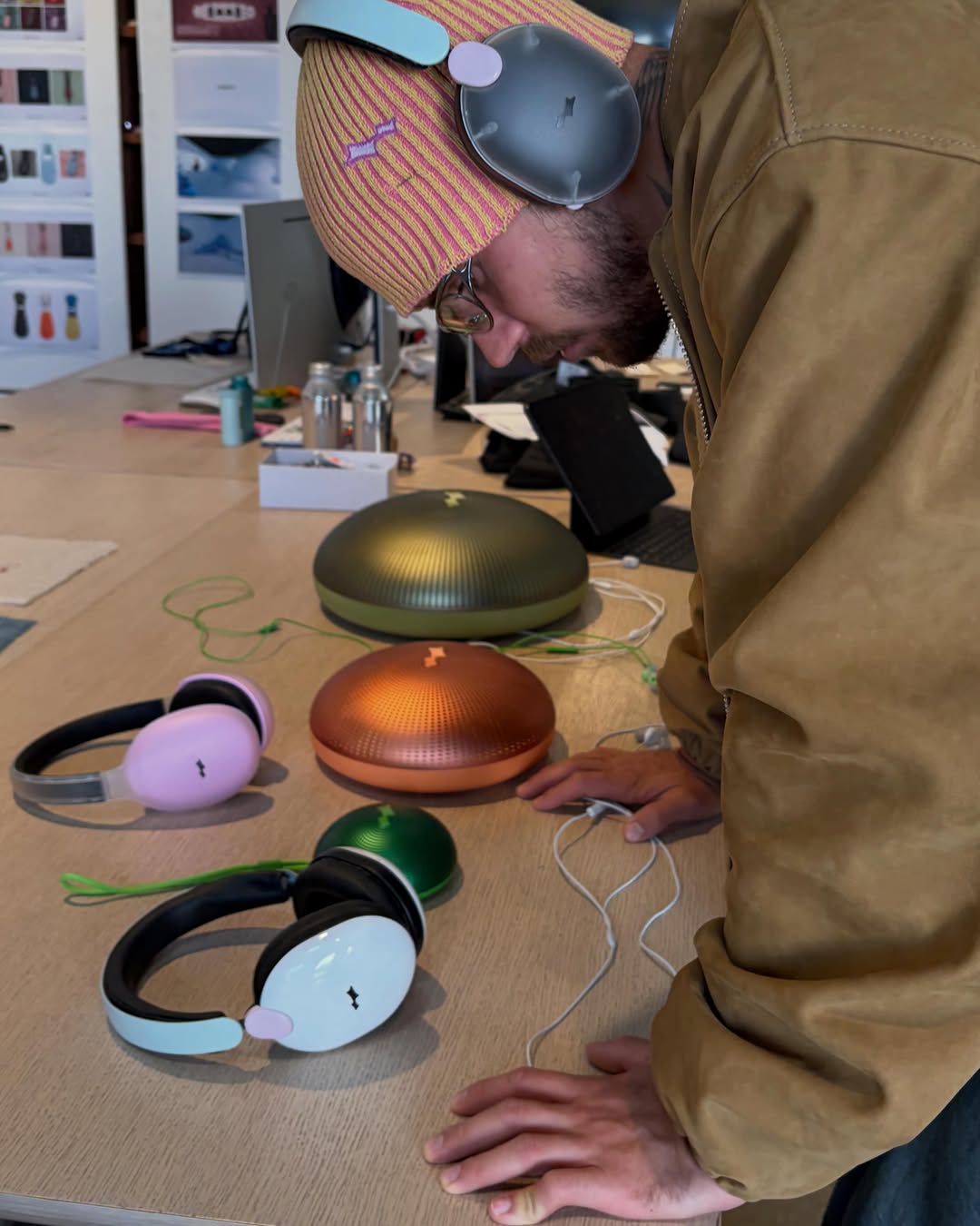 Person wearing pastel over-ear headphones and a striped beanie leans over a table examining a collection of colorful speakers and headphones in a design studio.