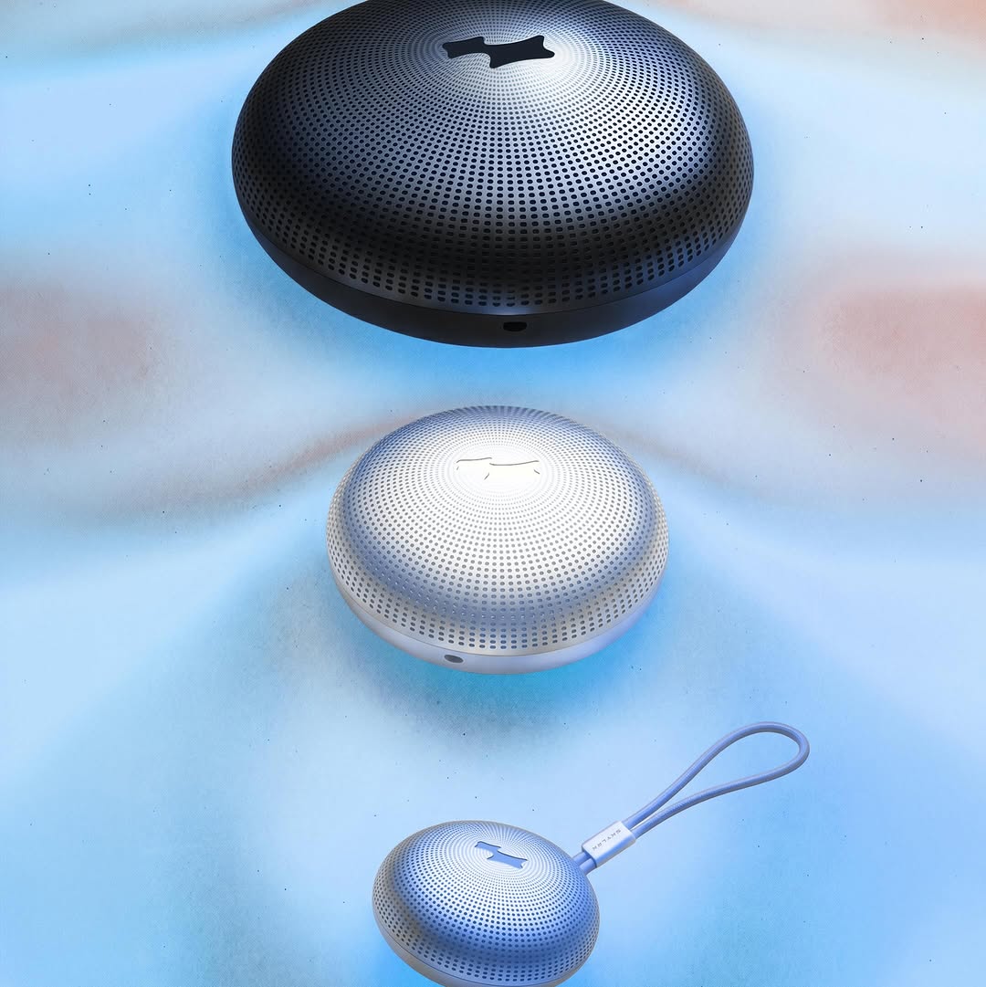 Three portable speakers in black, silver, and silver-with-lanyard configurations displayed against a cool blue gradient background.