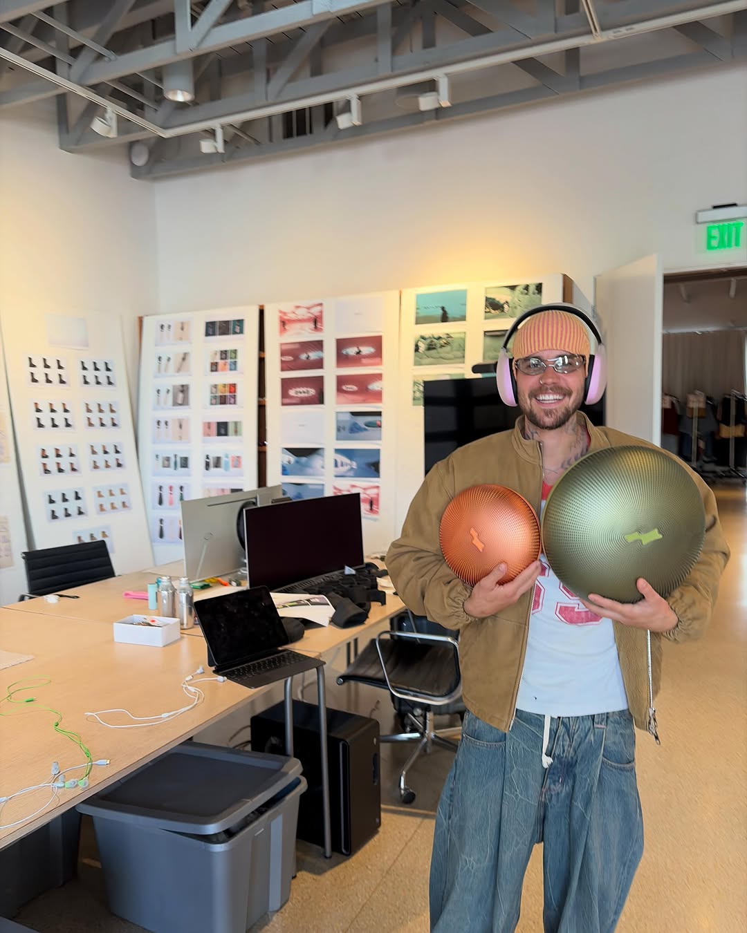 Smiling person wearing pink over-ear headphones holds two large portable speakers, one bronze and one olive green, in a modern office space.