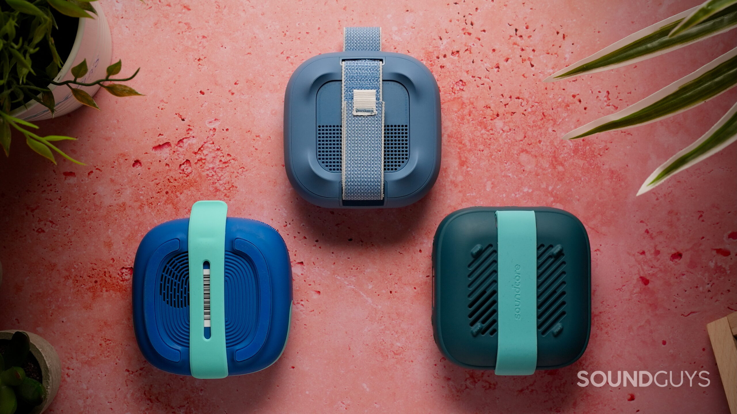 The rear of the Bose SoundLink Micro (2nd Gen), Tribit StormBox Micro 3, and Soundcore Boom Go 3i Bluetooth speakers showing their integrated carrying straps on a pink textured surface.