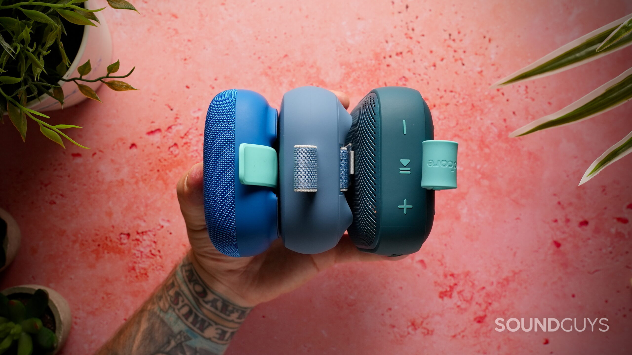 A hand holds the Tribit StormBox Micro 3, Bose SoundLink Micro (2nd Gen), and Soundcore Boom Go 3i side by side to compare their thickness.
