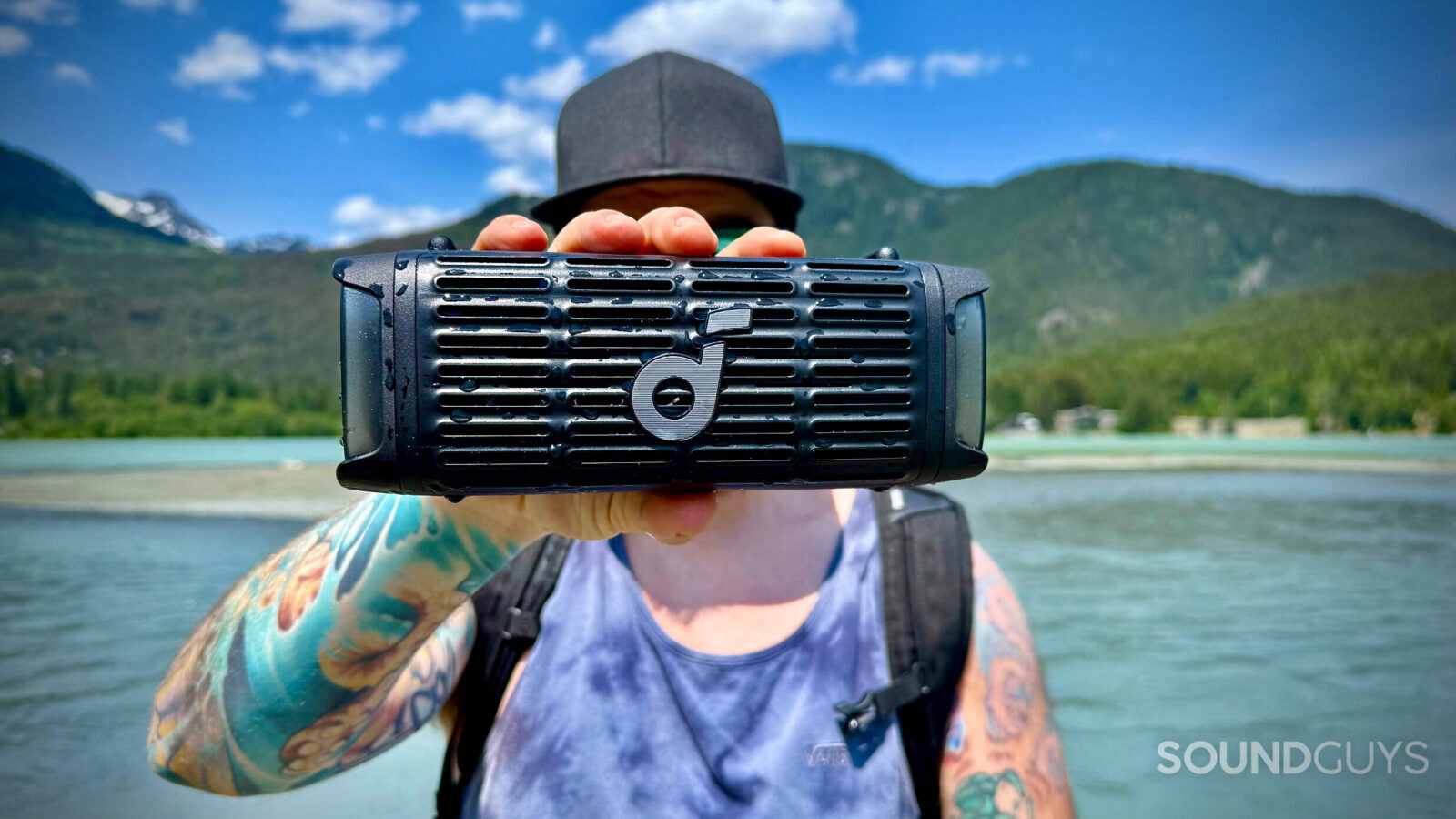 A man holding a Soundcore Boom 3i in front of his face with mountains and water in the background.