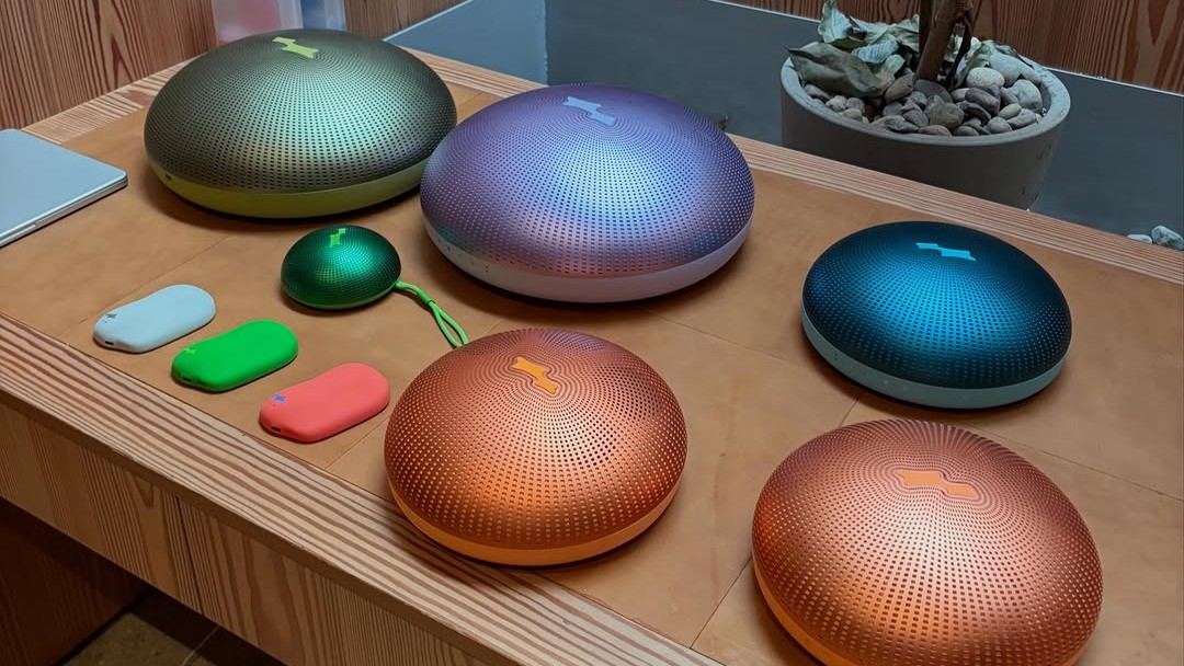 bieberspeakers Assortment of portable speakers and audio accessories in various colors displayed on a wooden table against a wood-paneled backdrop.