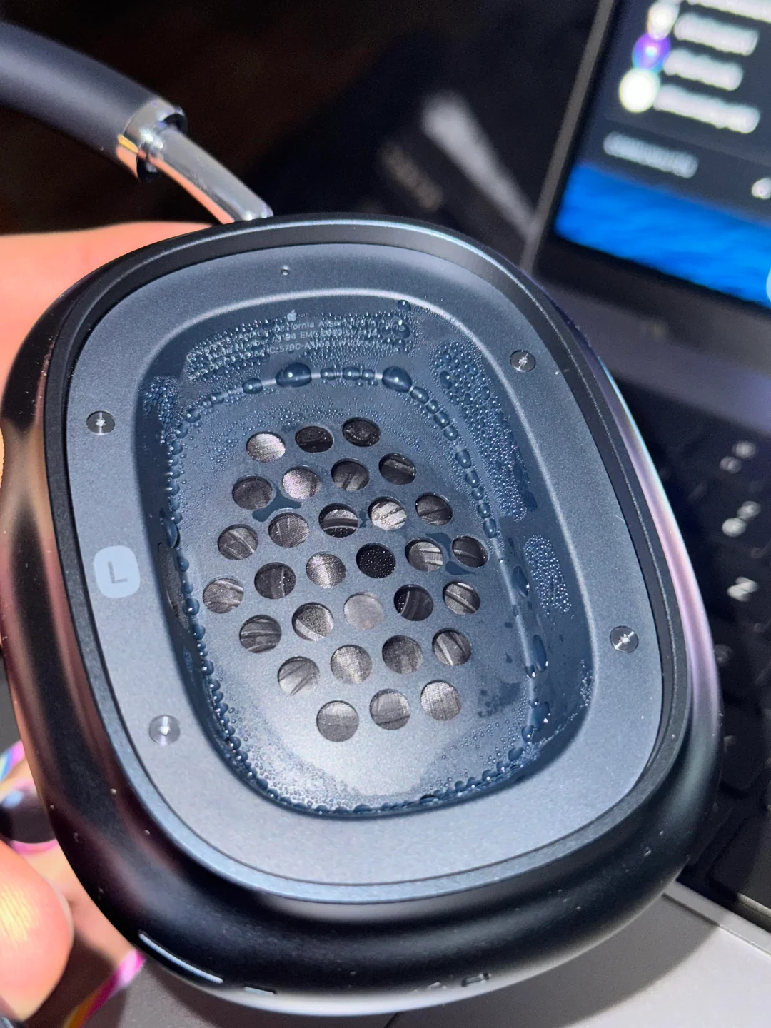 An Apple AirPods Max headphone held in hand with the ear cushion removed, revealing visible condensation and water droplets around the inside of the ear cup and speaker grille, a common effect caused by heat and humidity during use.