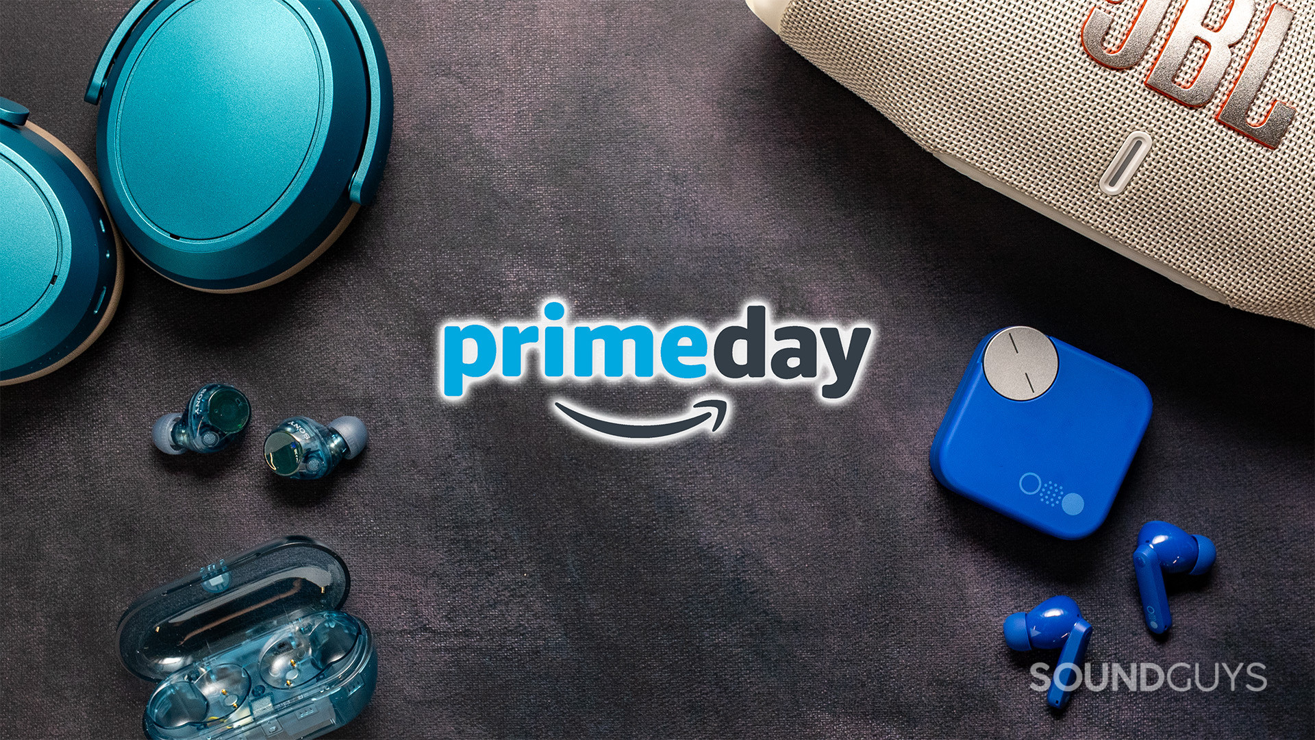 A flat-lay image featuring audio gear arranged around an Amazon Prime Day logo. Blue over-ear headphones sit in the upper left, a portable JBL speaker appears in the upper right, and two pairs of wireless earbuds with charging cases are positioned at the bottom left and right. The products surround the centered Prime Day branding on a dark textured background.