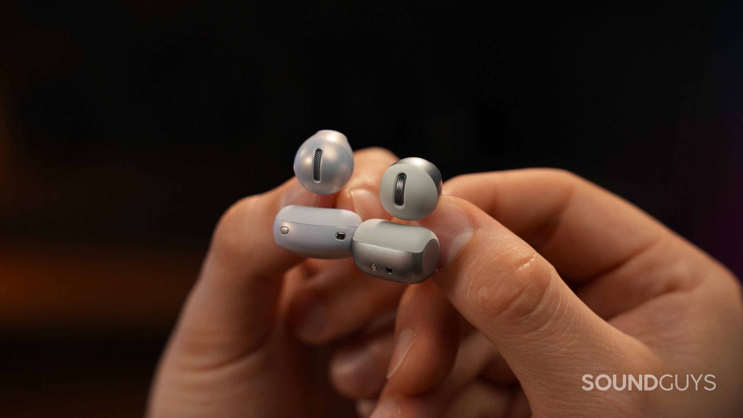 Close-up comparison of two clip-on open-ear earbuds held between fingers. The speaker grilles, microphones, and metallic accent finishes are visible, highlighting design differences between the two models.