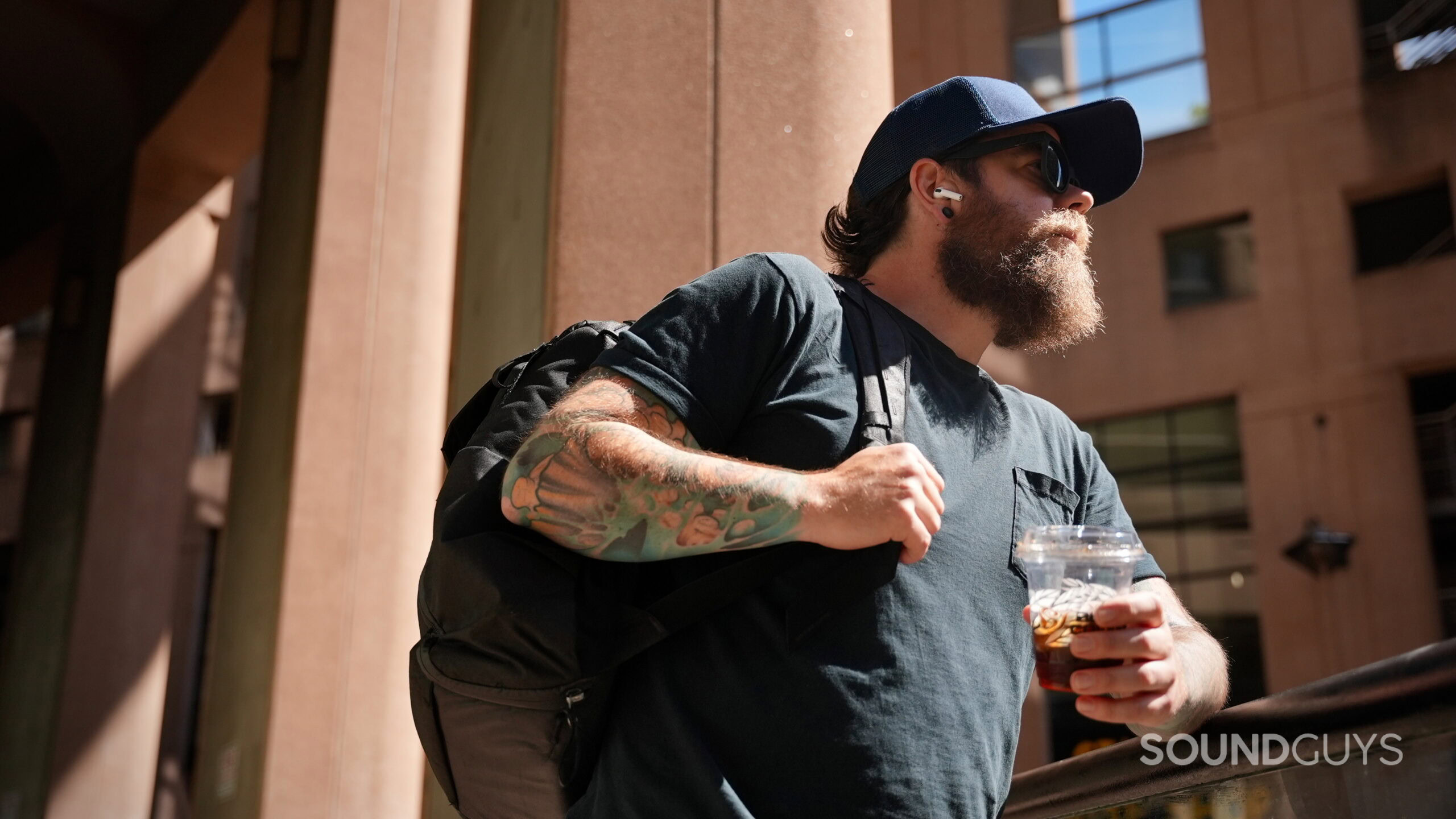 JBL Tour One and AirPods Pro 3 Travel feature - 1 (1) A person wearing AirPods Pro 3 earbuds while carrying a backpack and holding an iced coffee outdoors during a trip.