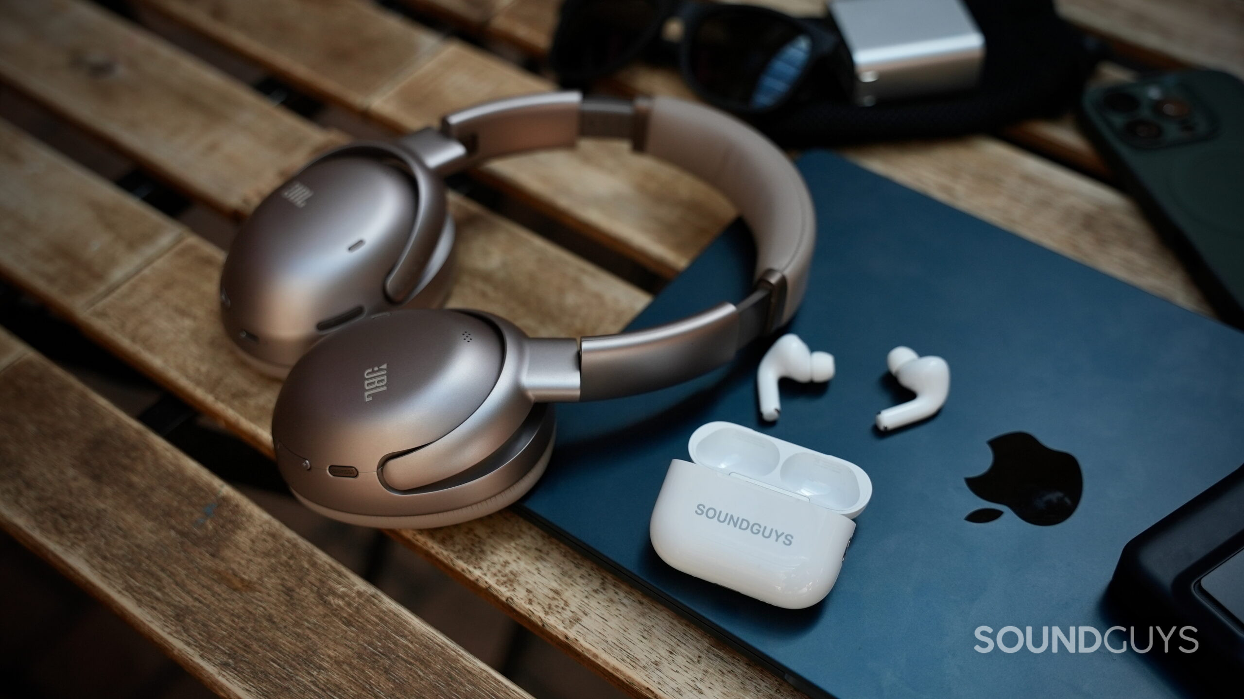 JBL Tour One and AirPods Pro 3 Travel feature - 2 The JBL Tour One M3 headphones and AirPods Pro 3 earbuds beside an iPad and travel accessories on a wooden table.