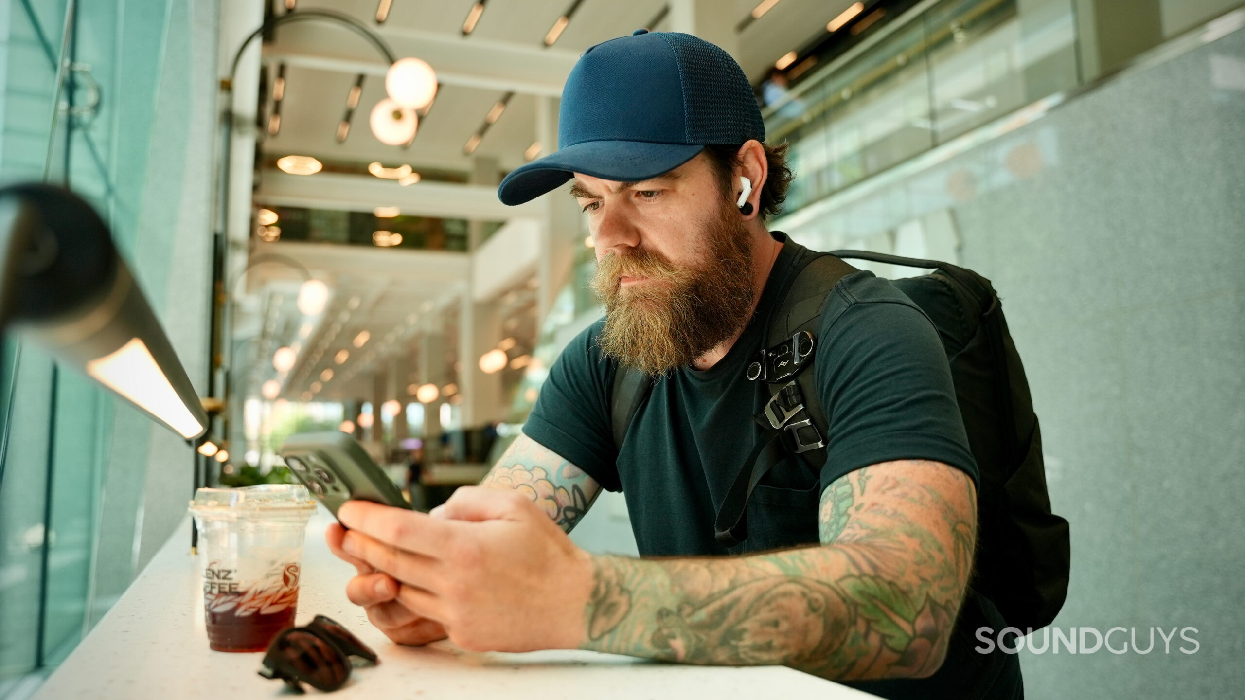 JBL Tour One and AirPods Pro 3 Travel feature - 3 A person wearing AirPods Pro 3 earbuds while using a smartphone with a backpack and iced coffee nearby.