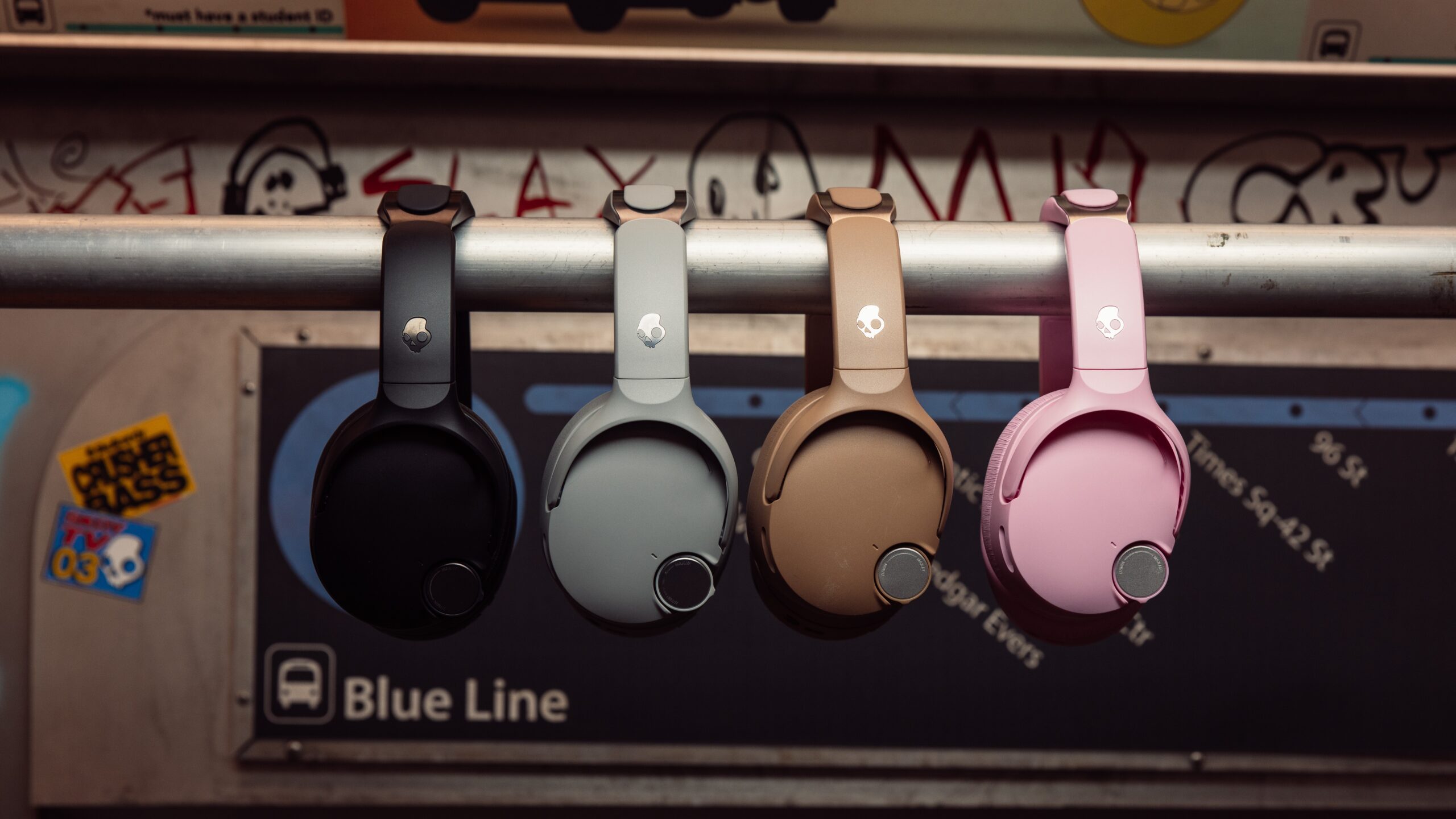Four Skullcandy Crusher 1080 ANC headphones in black, light gray, tan, and pink hang from a metal subway handrail above a Blue Line transit sign, showcasing the different colorways in an urban setting.