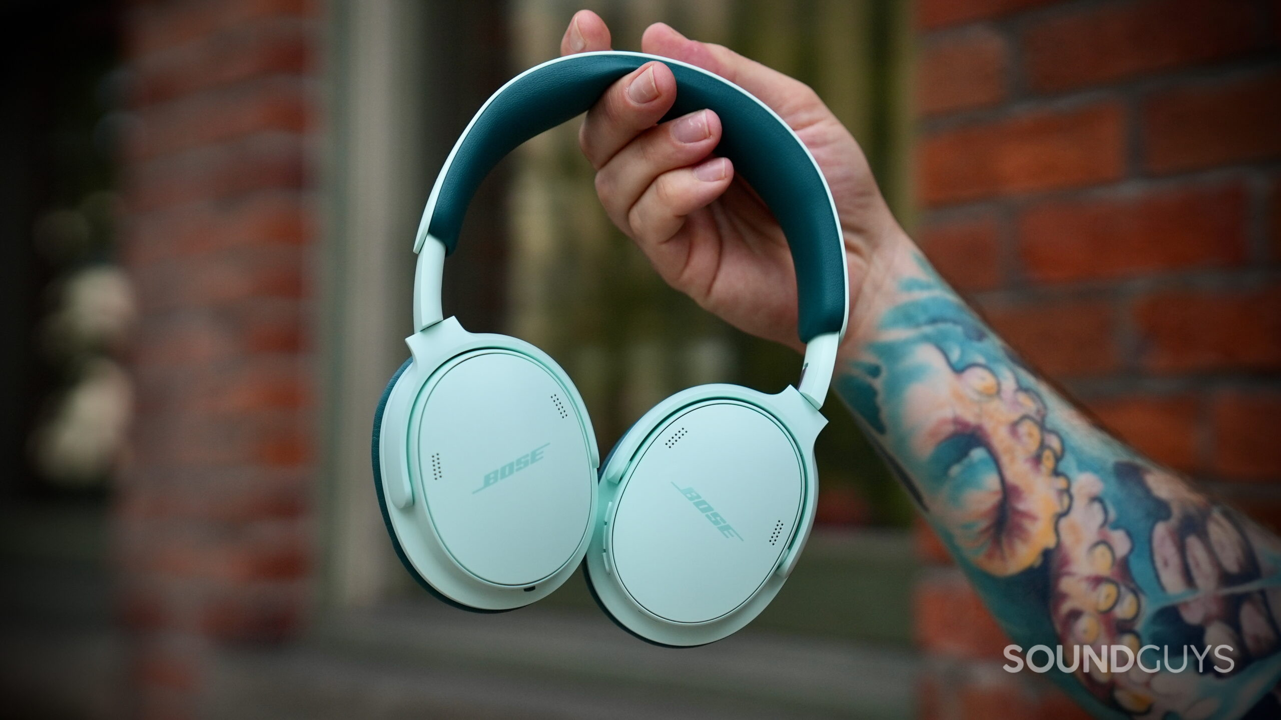 Bose QC Headphones (2nd Gen) - 2 The Bose QuietComfort Headphones (2nd Gen) held in one hand in front of a brick wall.