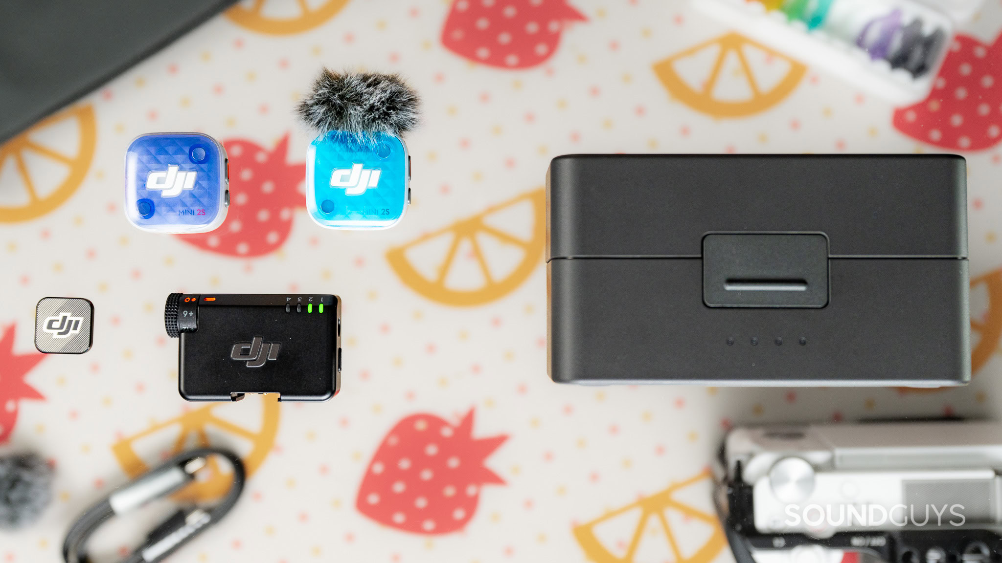 DJI Mic Mini 2S two transmitter top down with accessories Two wireless microphone transmitters, above a camera receiver, with a large case to fit both the receiver and transmitters, with some accessories in the background.