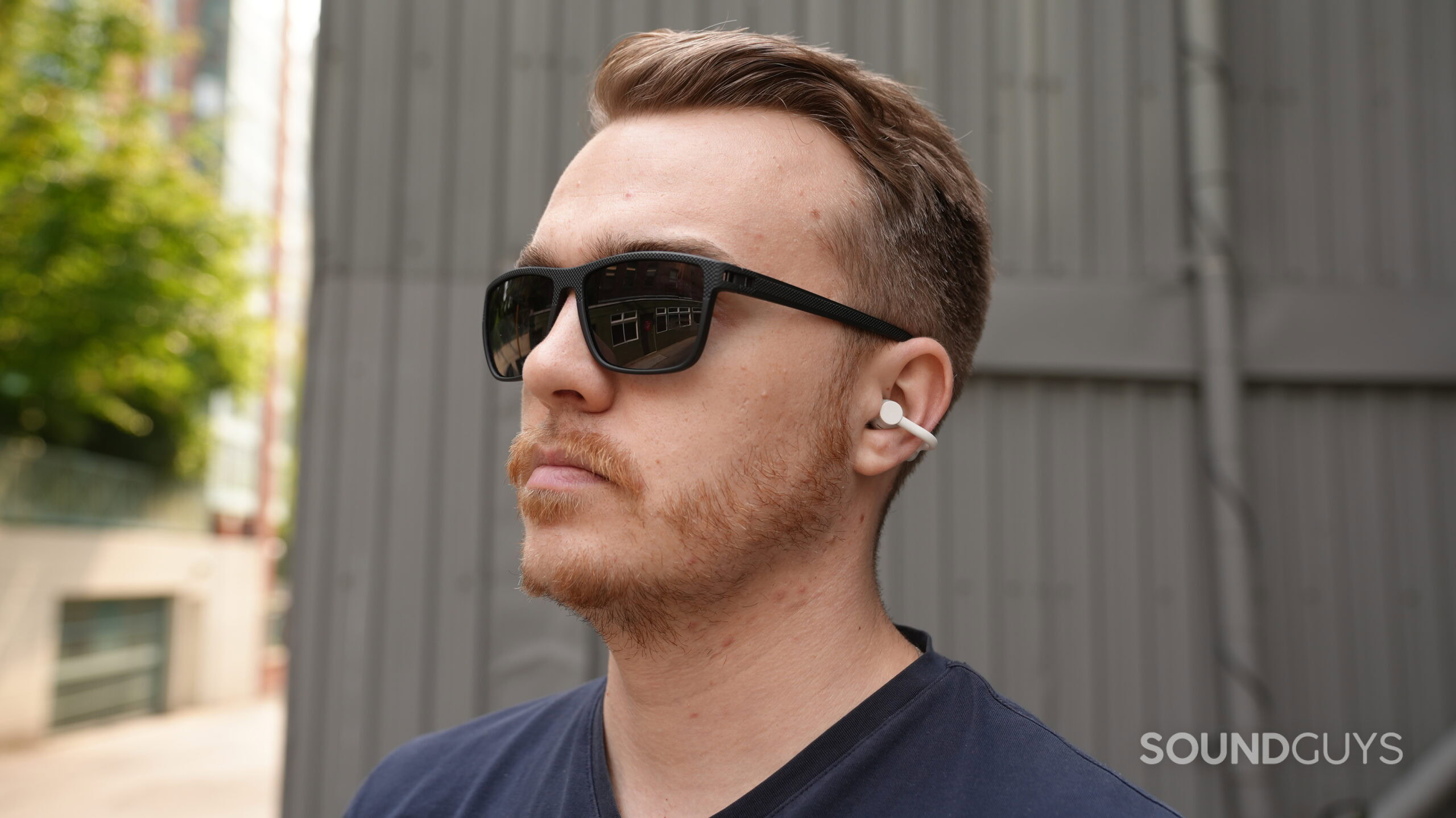 DSC00001 A man wearing sunglasses uses the white CMF Clip Pro open-ear earbuds outdoors. Caption: The CMF Clip Pro sits securely around the ear.