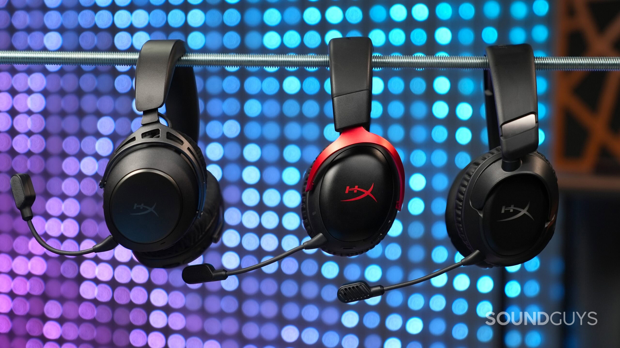 DSC00090 Three HyperX gaming headsets with boom microphones hanging from a metal rod against blurred purple and blue lights.