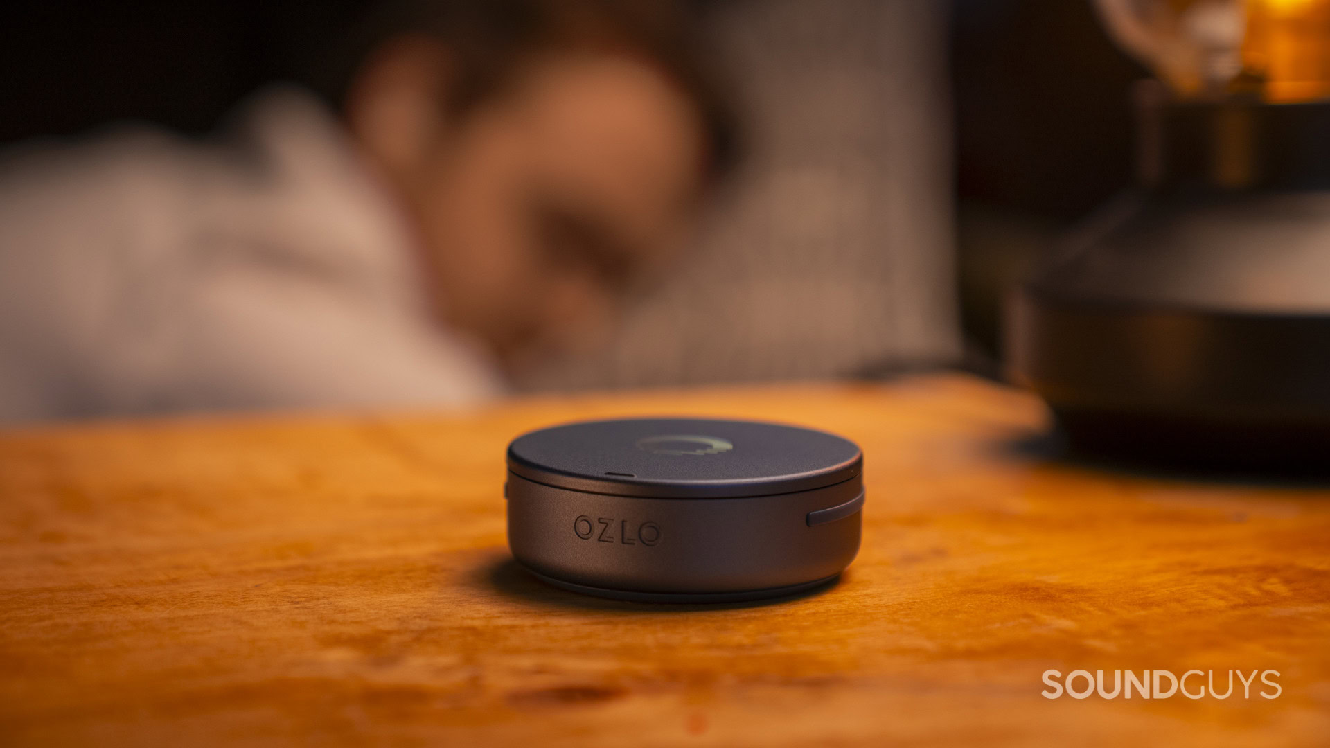 SG_Ozlo Sleepbuds 2-3 Ozlo Sleepbuds 2 charging case on a bedside table beside a sleeping person.