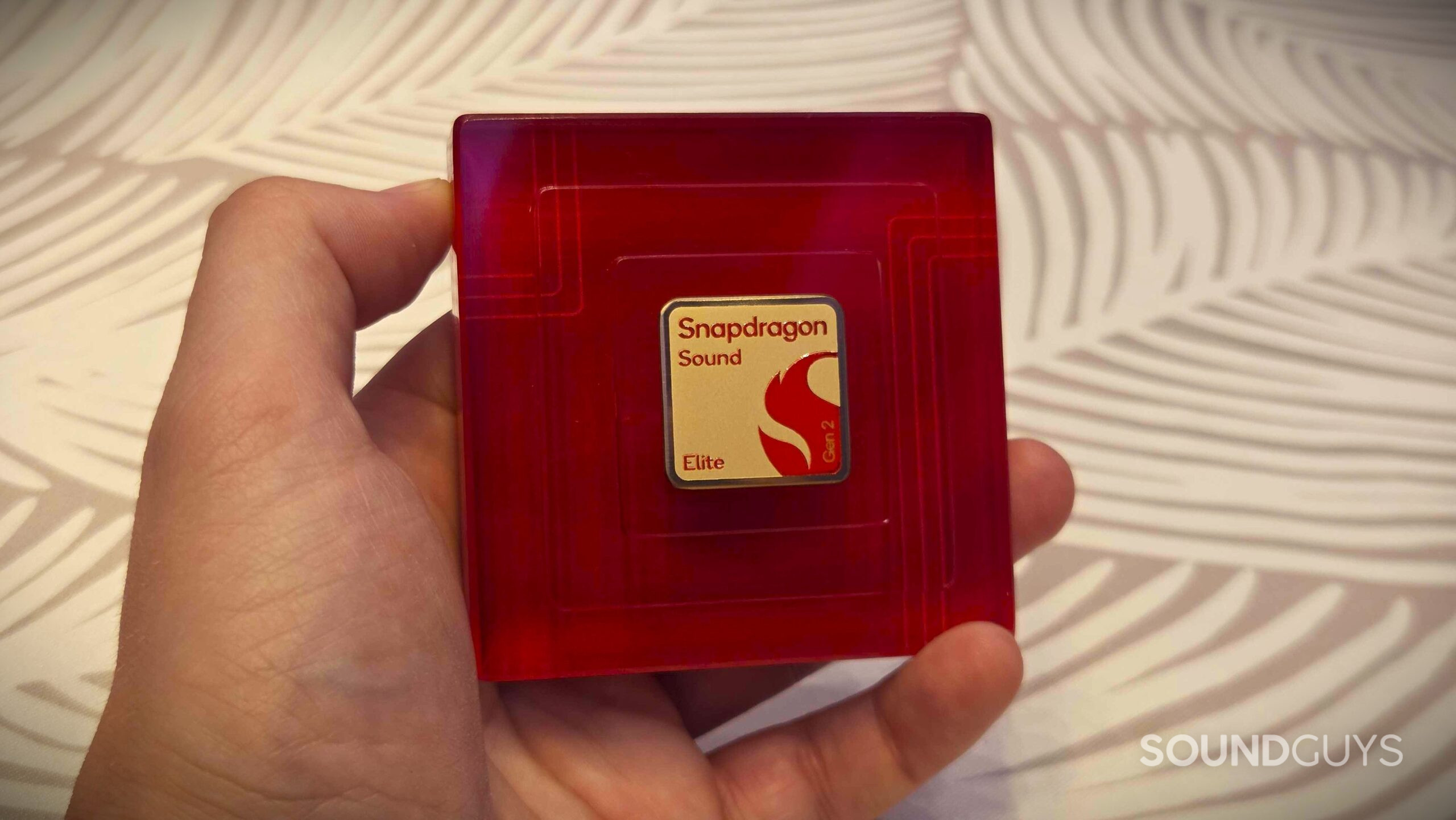 1000053167 A hand holds a red display piece with a gold Snapdragon Sound Elite Gen 2 badge against a patterned wall.