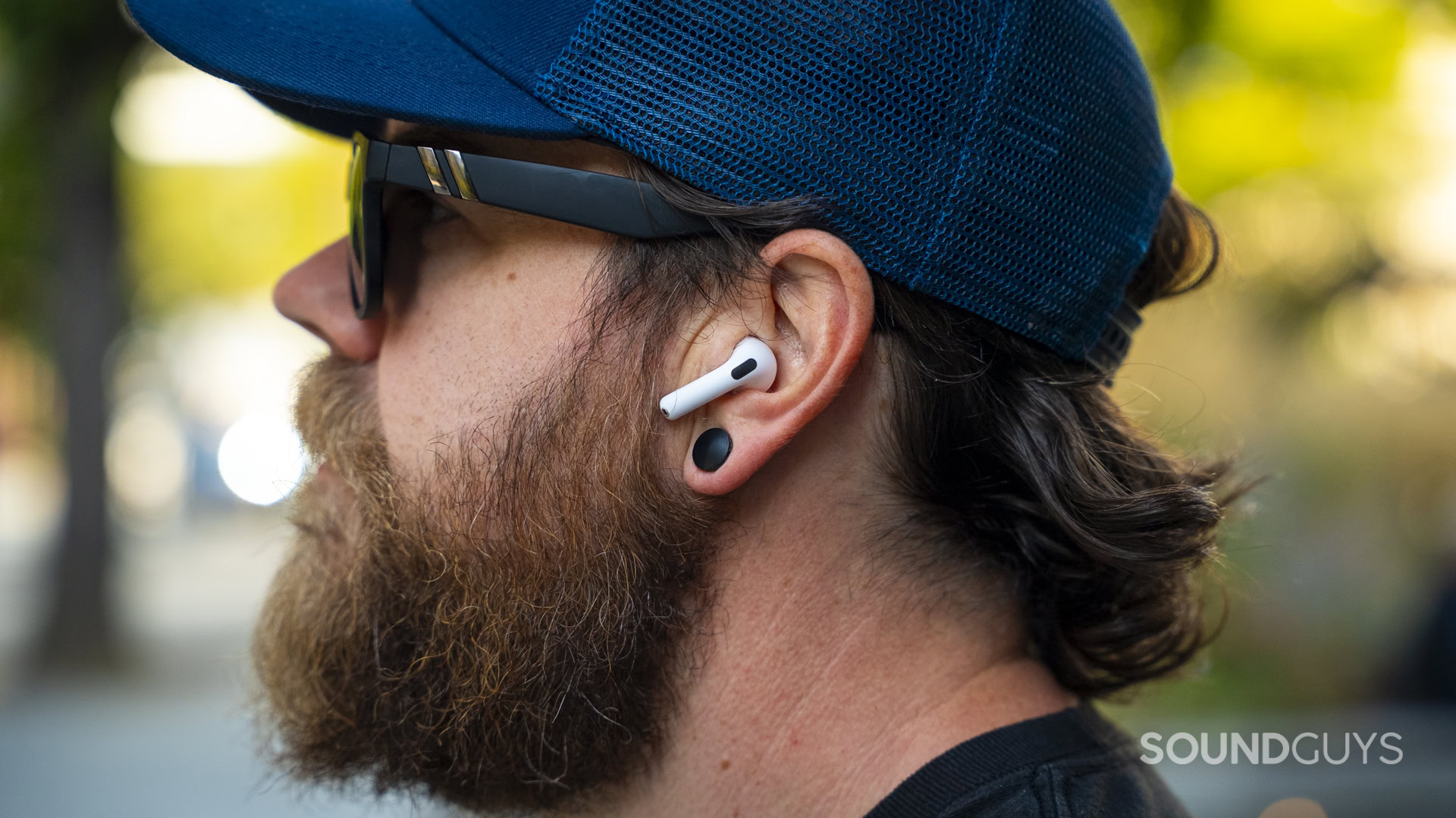 Apple AirPods 5 and Apple AirPods 5 with Wireless Charging-01 A man wearing a white Apple AirPod outdoors with sunglasses and a blue cap.