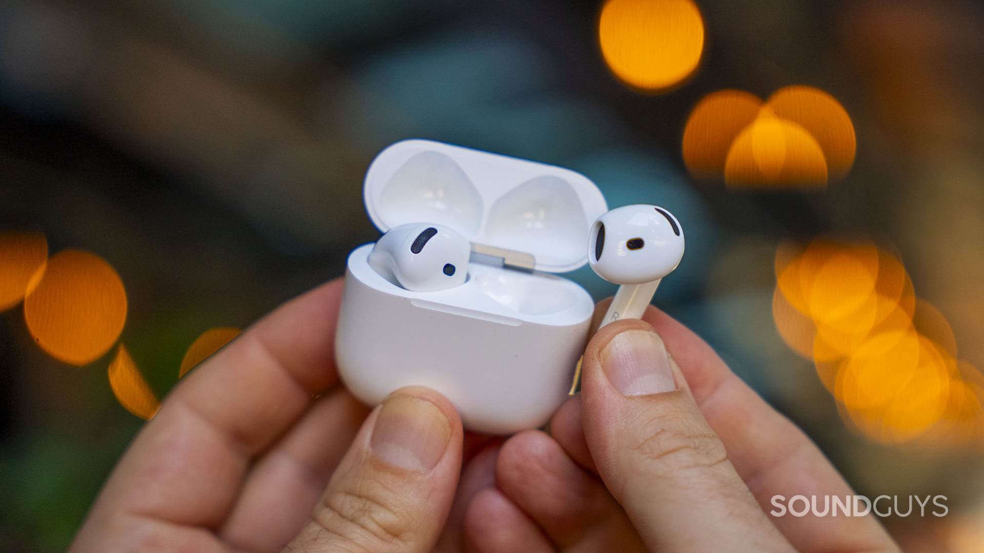 Apple AirPods 5 and Apple AirPods 5 with Wireless Charging-22 One Apple AirPod held beside its open charging case against warm background lights.