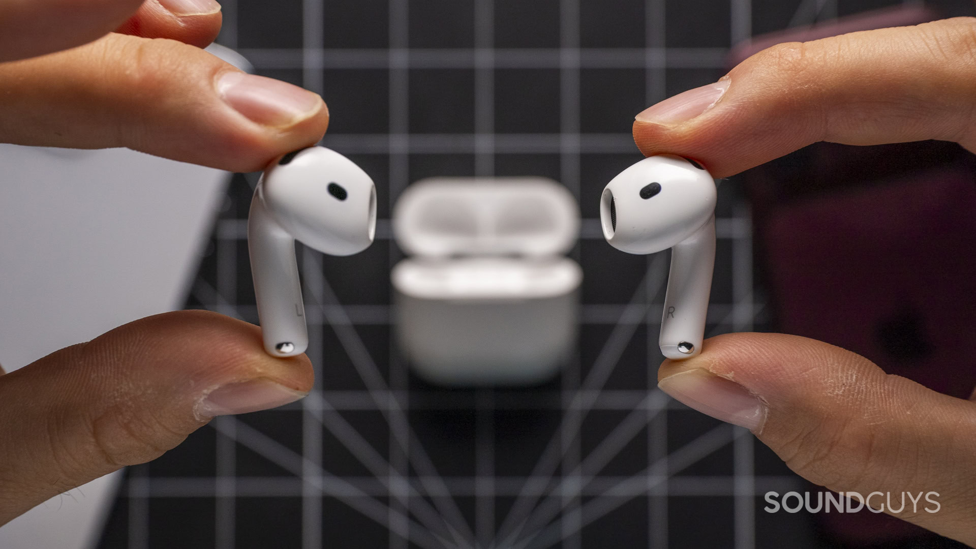 Apple AirPods 5 and Apple AirPods 5 with Wireless Charging-23 A pair of white Apple AirPods held between two hands with the charging case behind them.