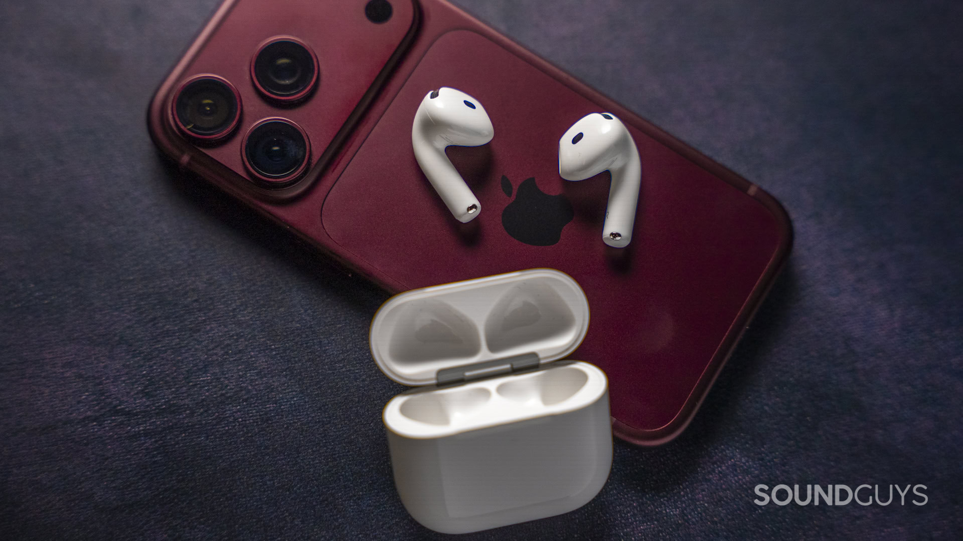 Apple AirPods 5 and Apple AirPods 5 with Wireless Charging-30 White Apple AirPods and their open charging case arranged on a red iPhone.
