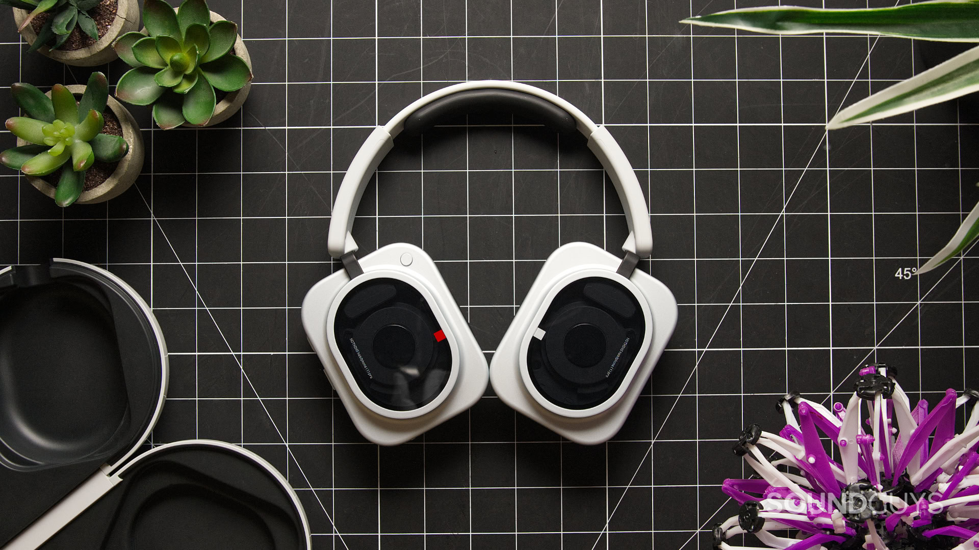 A photo of the Nothing Headphone (1) Pro sitting atop a table with plants, its open case, and a toy.