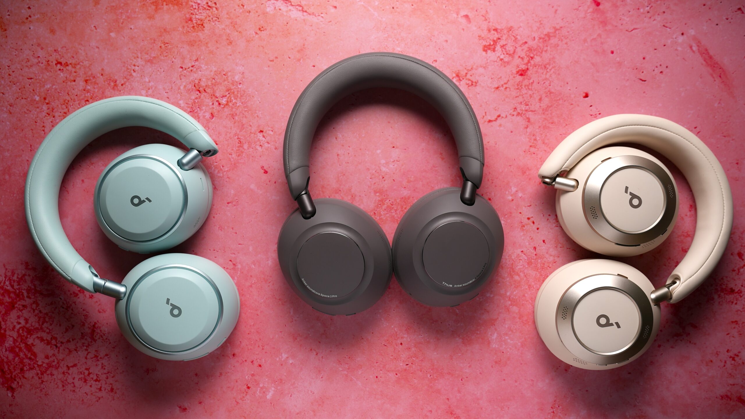 soundcore space 2 pro with space 2 and space oen pro headphones Three pairs of Soundcore over-ear headphones in blue, gray, and white arranged on a pink textured surface.