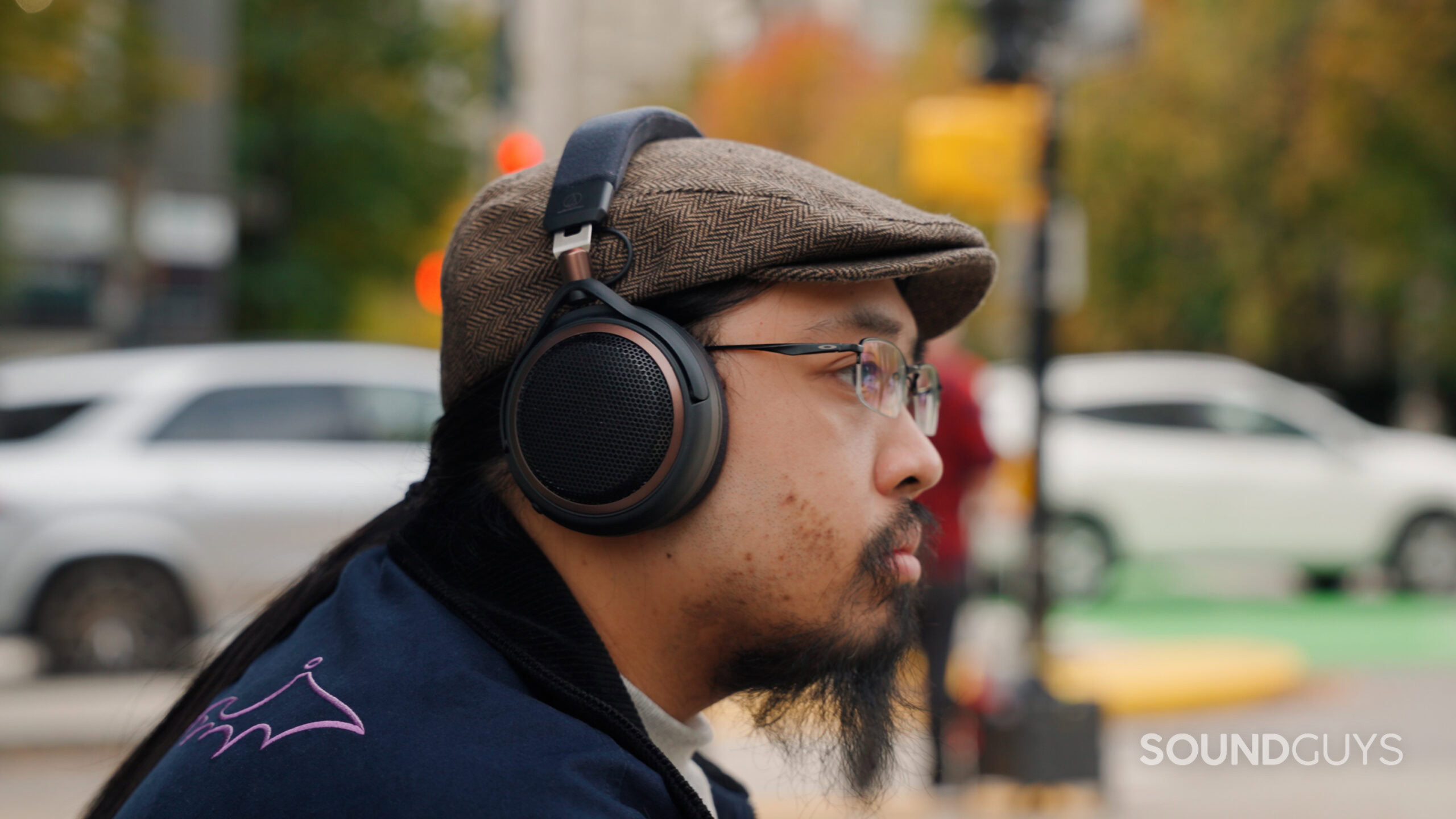 Audio-Technica ATH-AL5NC worn on street A side-profile photo of a person wearing black over-ear Audio-Technica headphones, a brown herringbone flat cap, rectangular glasses, and a navy jacket with purple embroidery. White cars and colorful city lights appear softly blurred in the urban background.