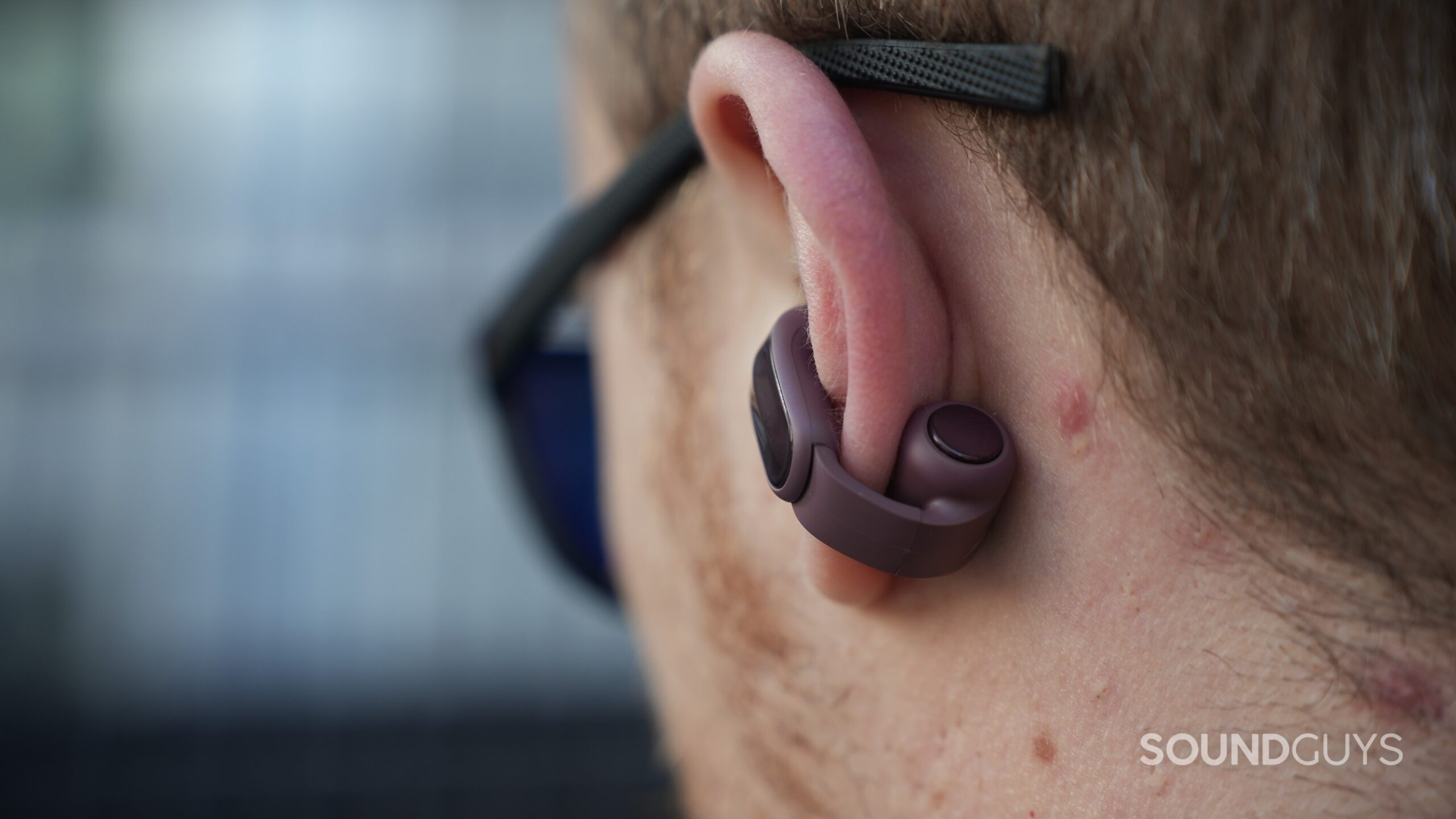 DSC09917 Rear close-up of a Cherry Chocolate Bose Ultra Open 2 earbud wrapped around an ear, showing its round control button beneath sunglasses.